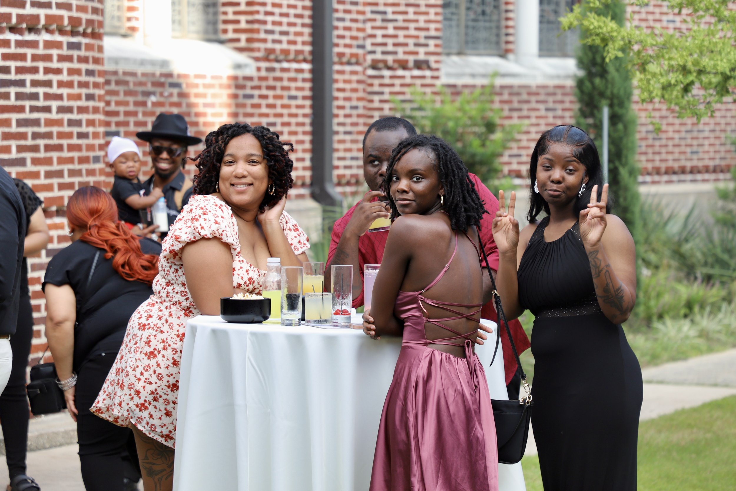 Group of friends standing around a white high-top table outdoors at a social event, some smiling and making peace signs, with drinks on the table, brick building, and greenery in the background.