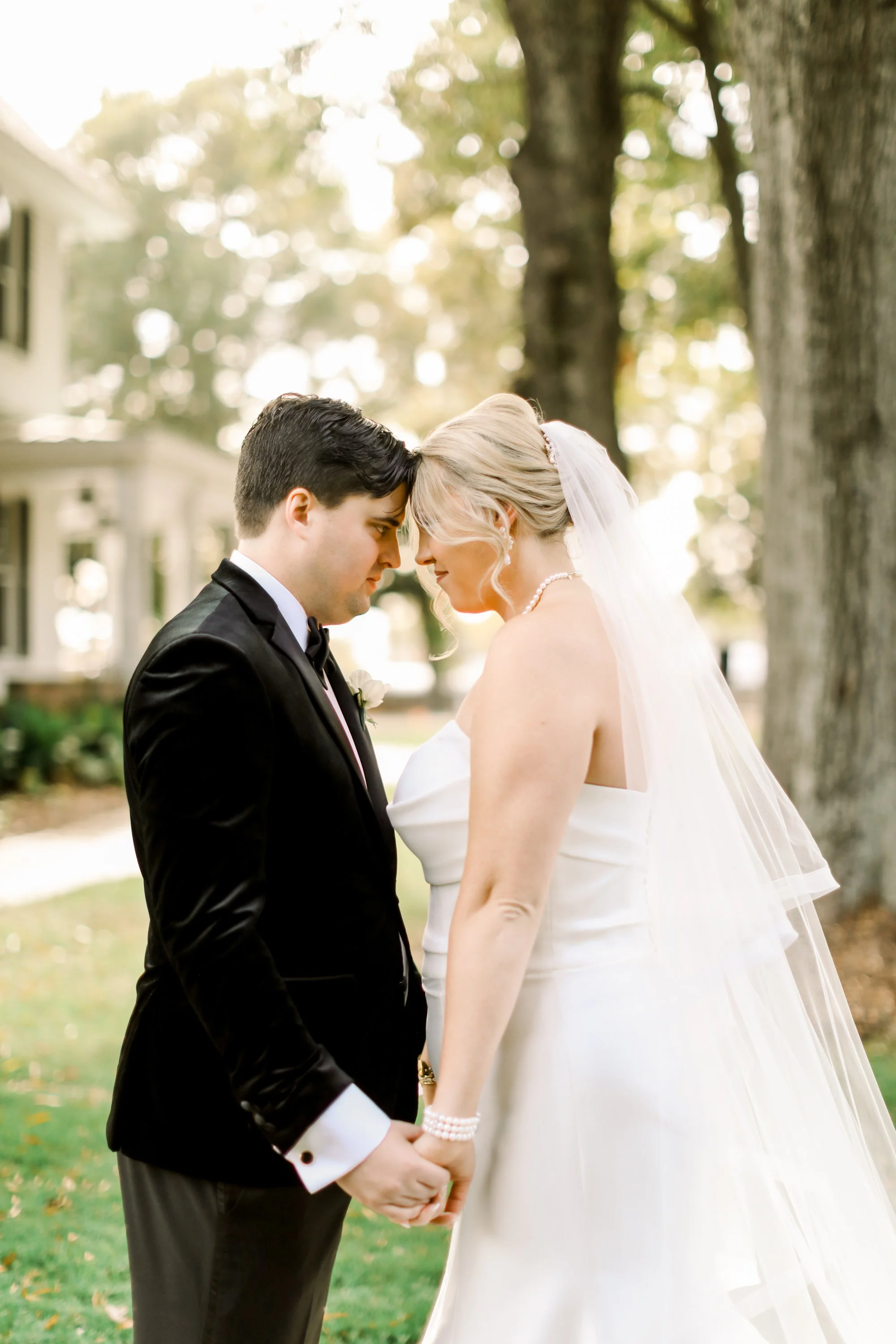A bride and groom in wedding attire holding hands and leaning foreheads together outdoors under trees