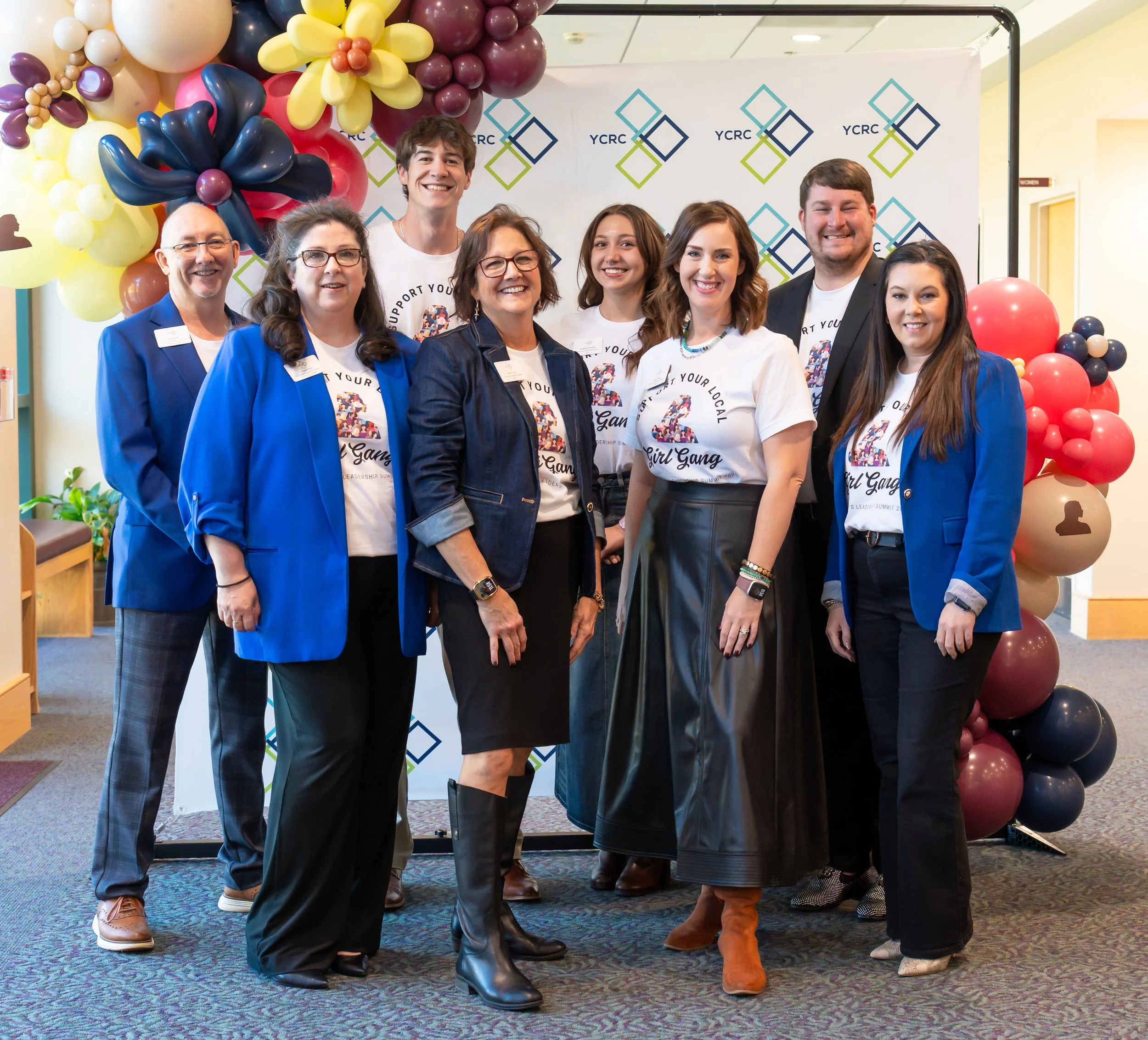 Group of nine people standing together in front of a backdrop with a colorful balloon arrangement on one side. They are all smiling and wearing casual or semi-formal attire, with some in matching T-shirts and others in blue blazers. The setting appea