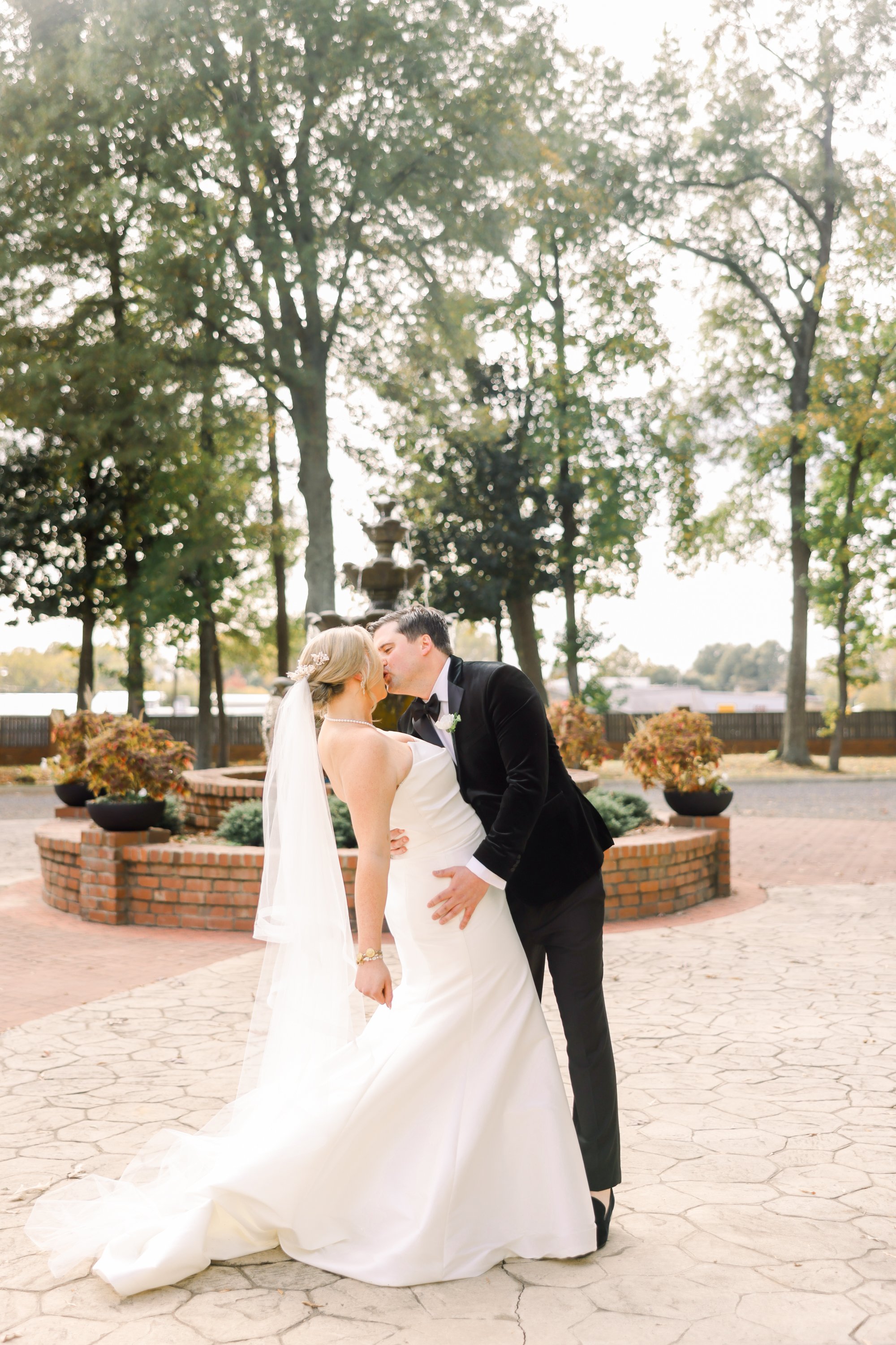 A bride and groom share a kiss outdoors in a park with trees, a fountain in the background, and a brick pathway.