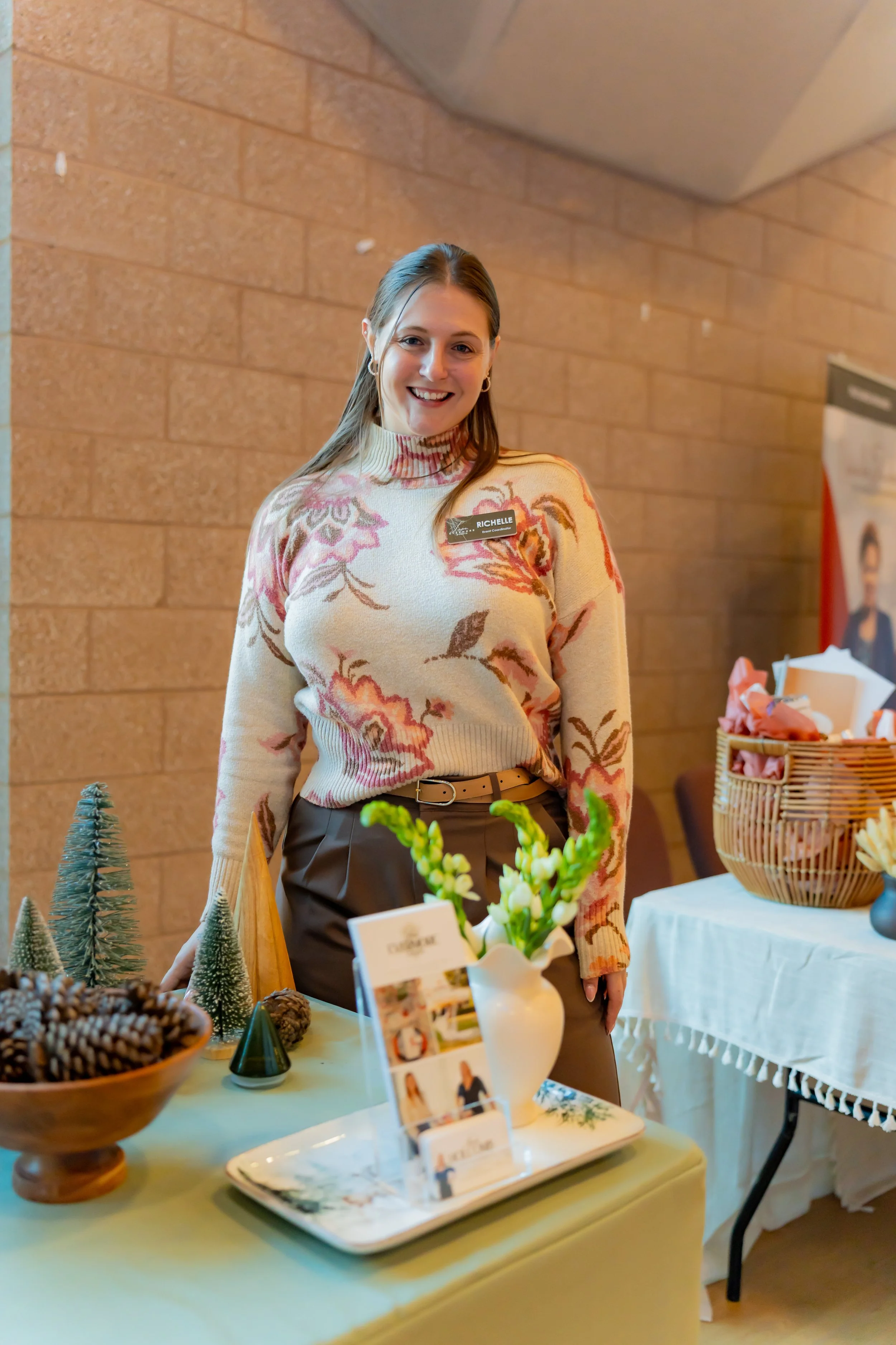 A smiling woman wearing a patterned sweater and brown pants standing behind a table with holiday decorations, including pinecones, small Christmas trees, and a vase with white flowers, inside a room with brick walls.