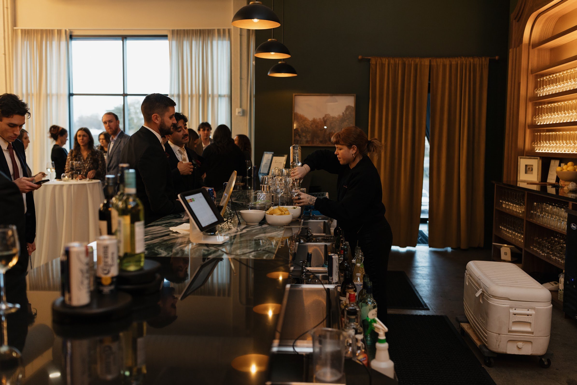People at a bar with a bartender preparing drinks, in a room with large windows, hanging lights, and curtains.