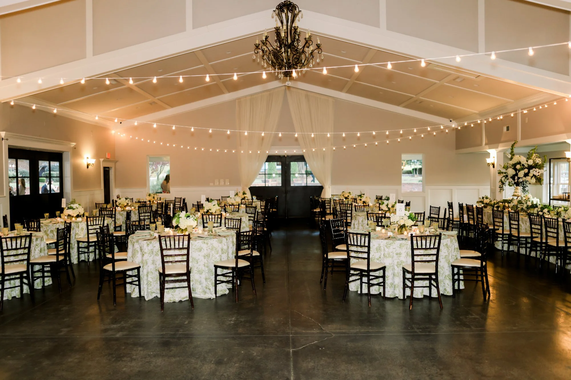 Elegant event hall decorated for a celebration with round tables covered in floral tablecloths, floral centerpieces, string lights overhead, and a chandelier hanging from the ceiling.