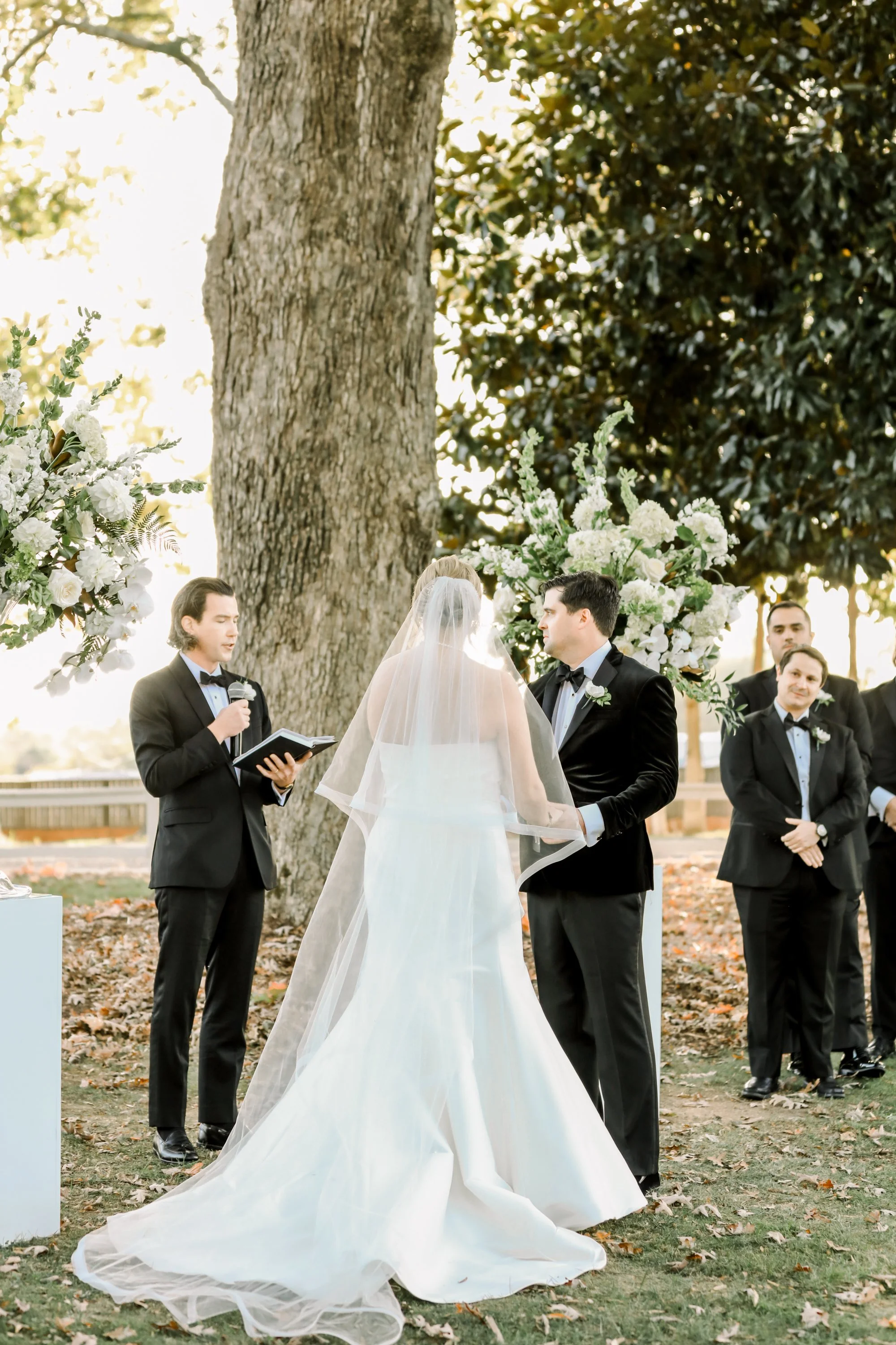 Bride and groom exchanging vows outdoors beneath large trees, with officiant holding a book and a floral arch nearby.
