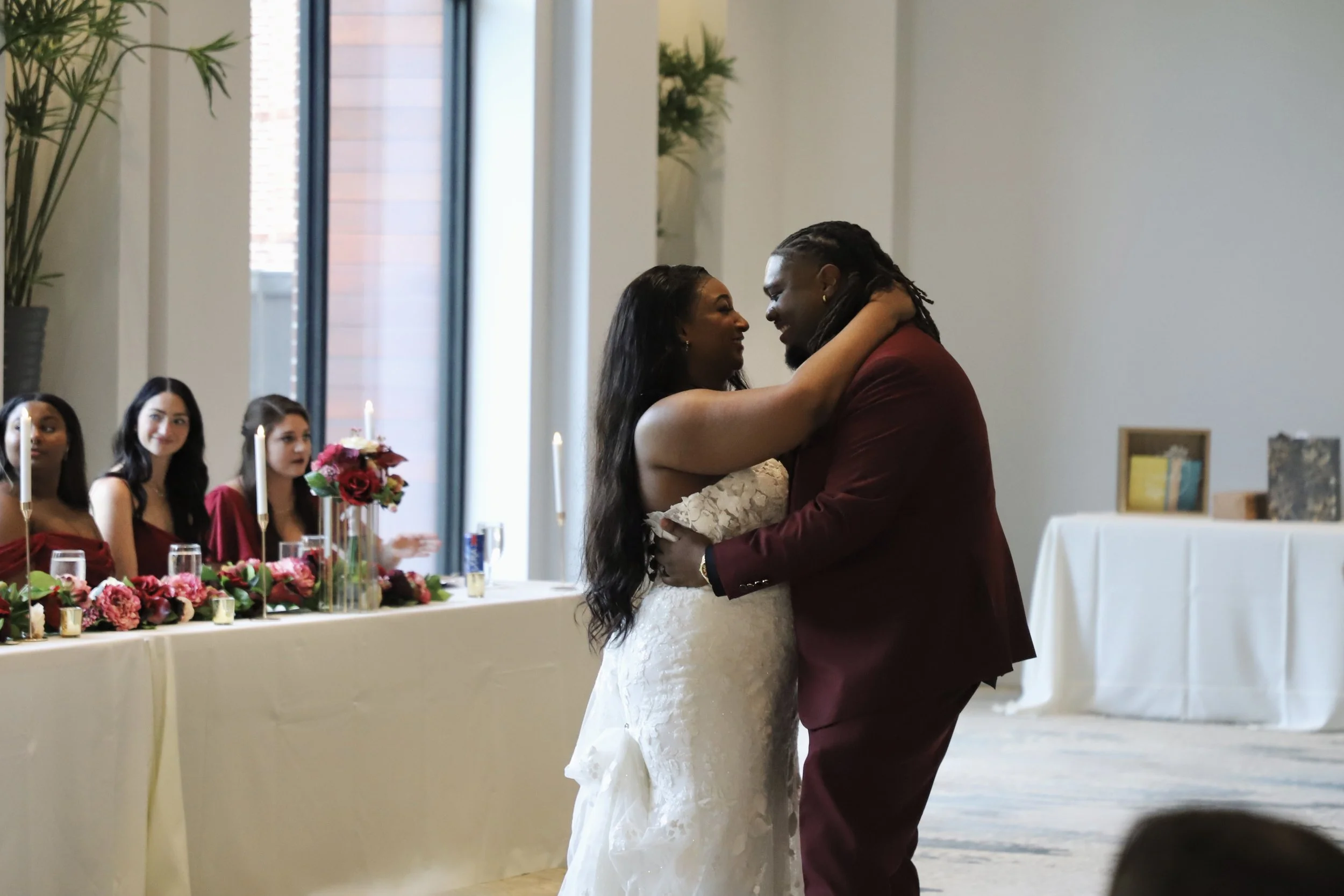 A bride and groom dancing at their wedding reception with three bridesmaids sitting at a decorated table in the background.