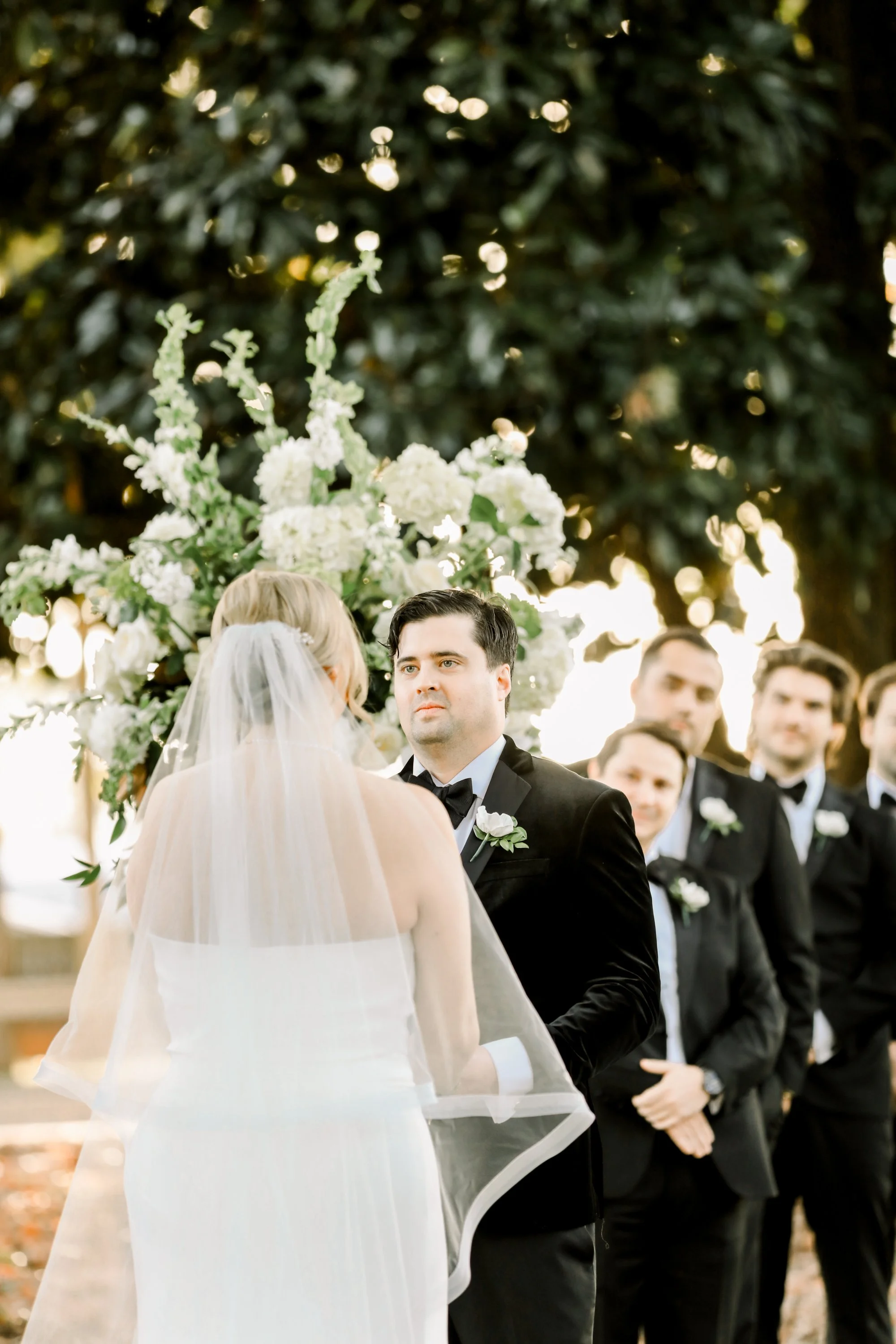 A bride and groom wedded vows during an outdoor wedding ceremony with green trees and a floral arch in the background, along with groomsmen in black tuxedos.
