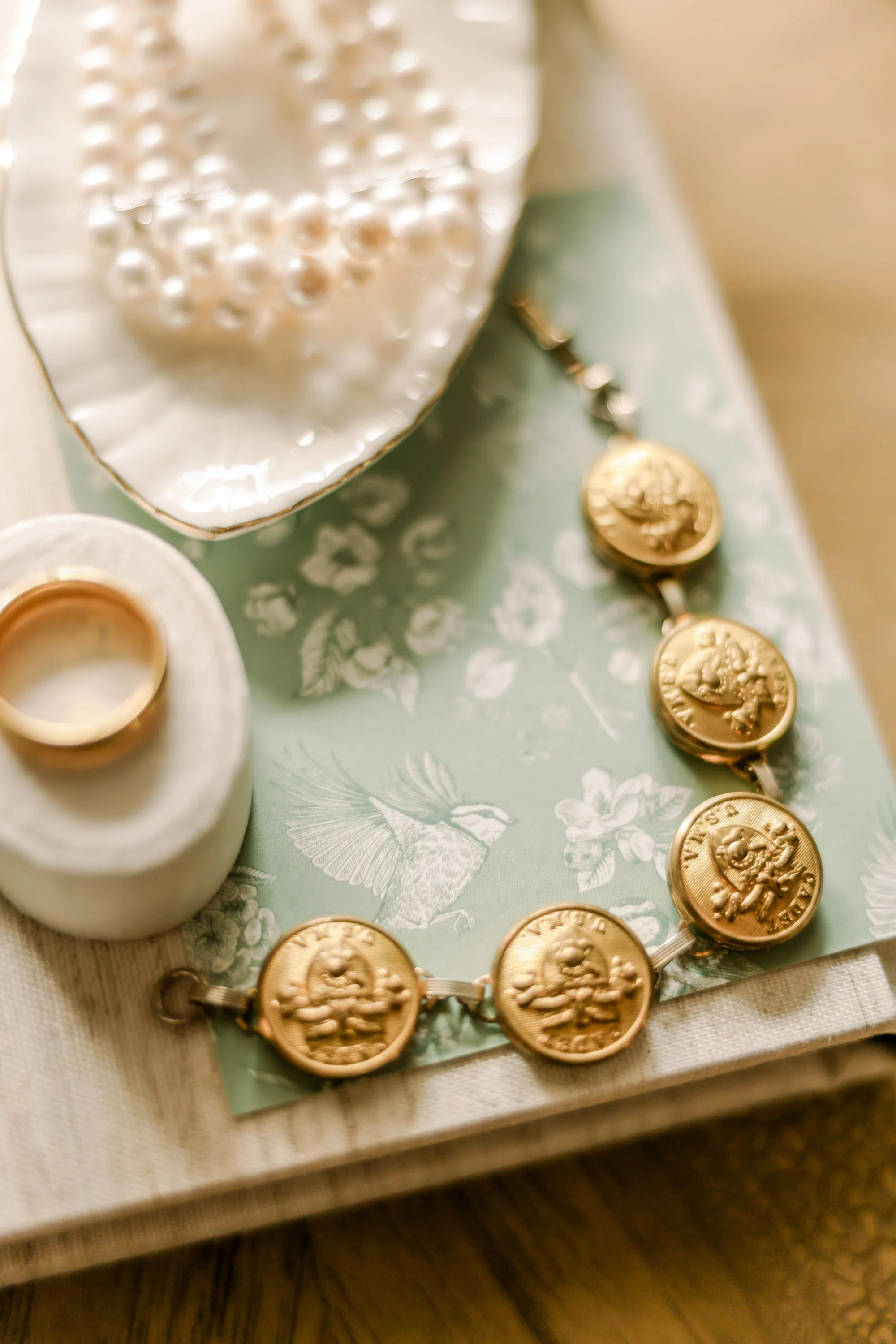 Gold coin bracelet, pearl necklace, gold ring, and decorative papers with floral and bird patterns on a textured surface.