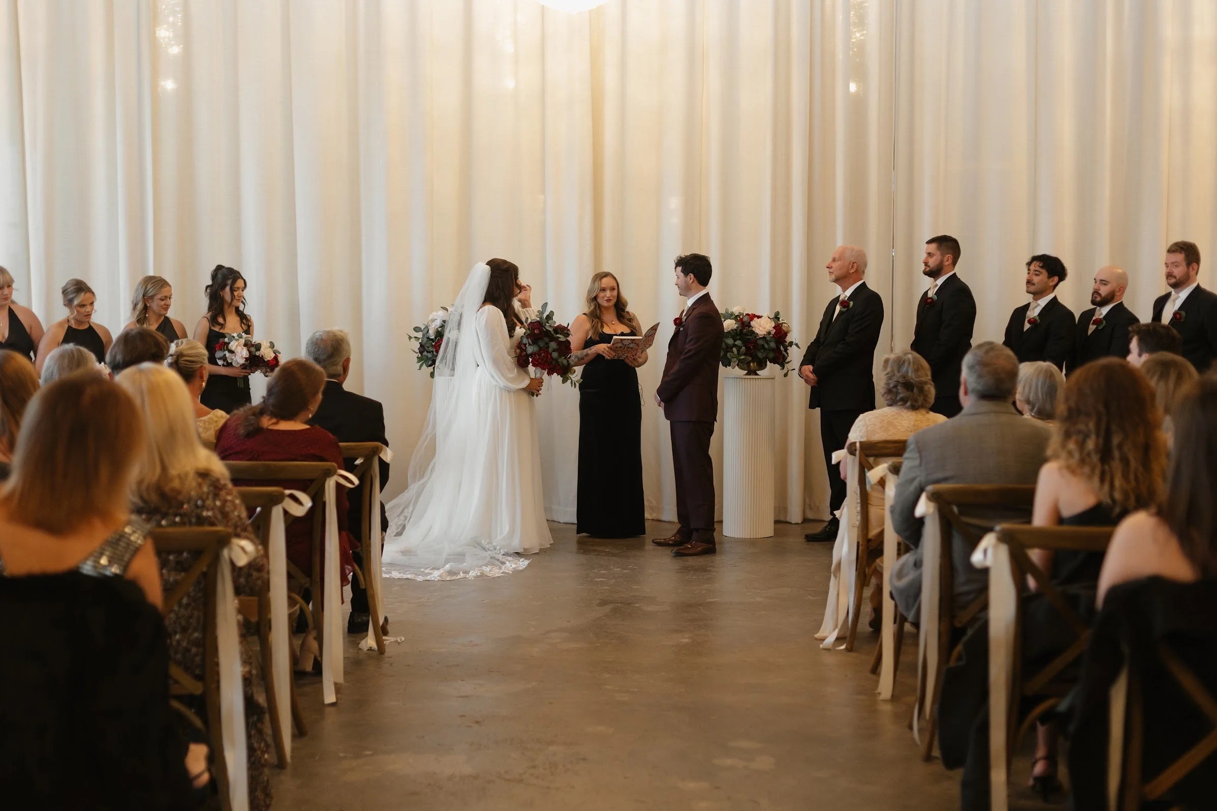 Bride and groom exchanging vows during a wedding ceremony with bridesmaids and groomsmen standing on either side, seated guests in the audience.