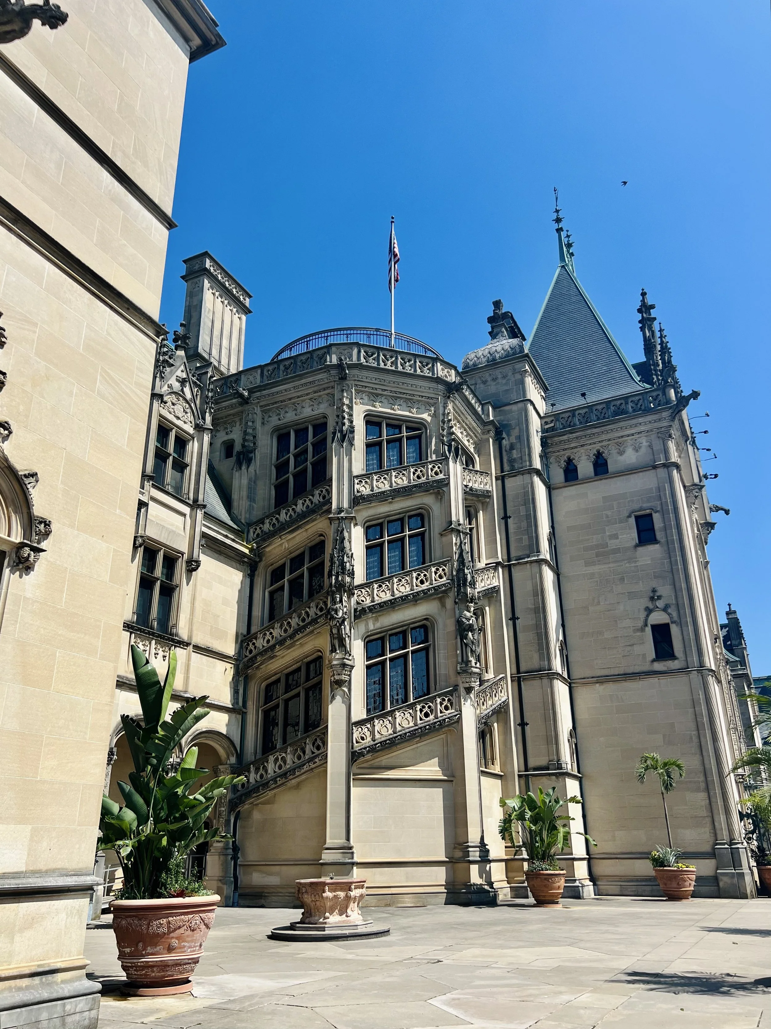 A historic castle-like building with ornate architectural details, surrounded by potted plants, under a clear blue sky.