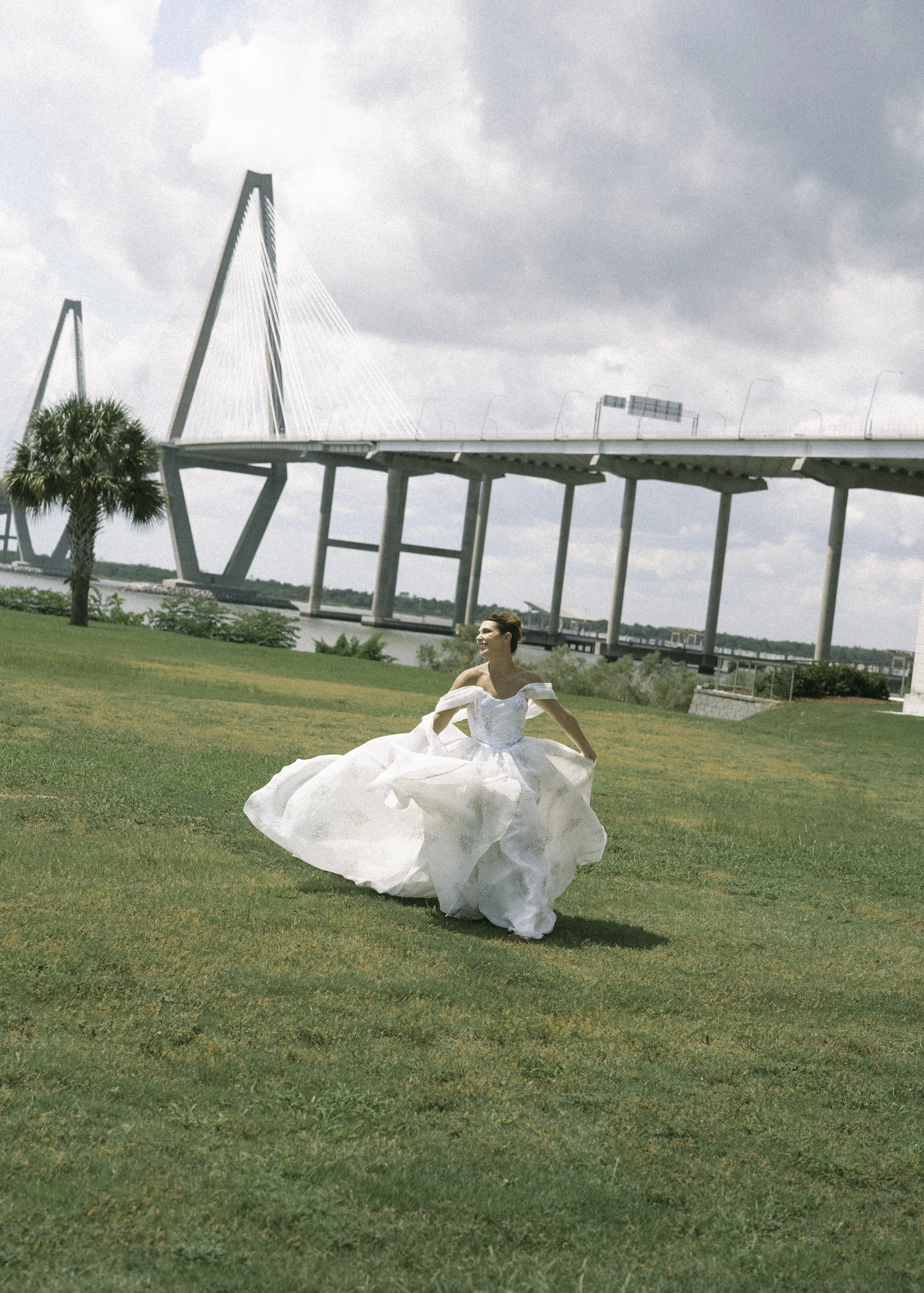 A woman in a white wedding dress standing on a grassy area, with a large bridge in the background.