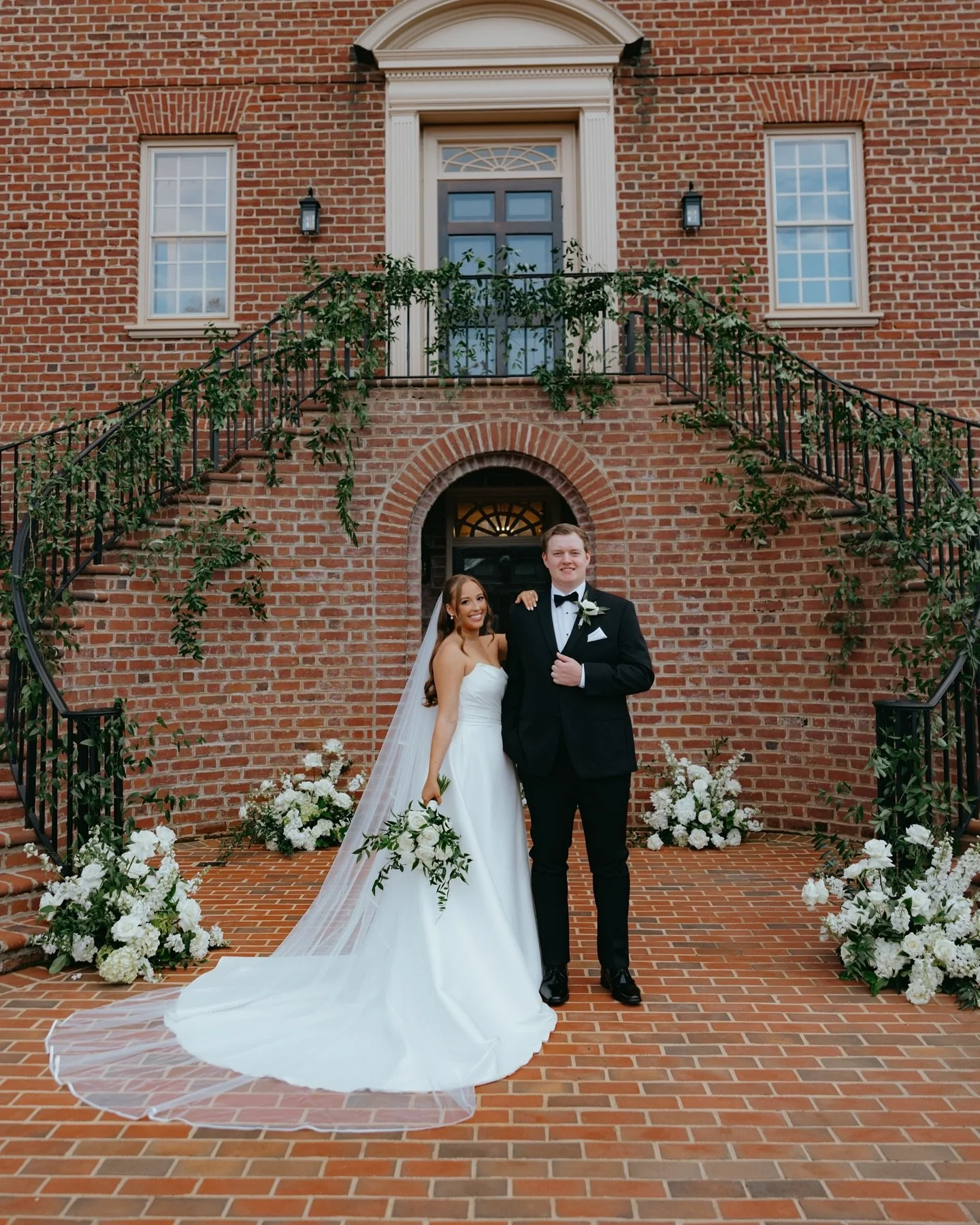 A bride and groom standing on brick steps outside a brick building, smiling for a wedding photo. The bride is in a white gown with a long veil and bouquet, the groom in a black tuxedo with a bow tie. Floral arrangements are on either side of them.
