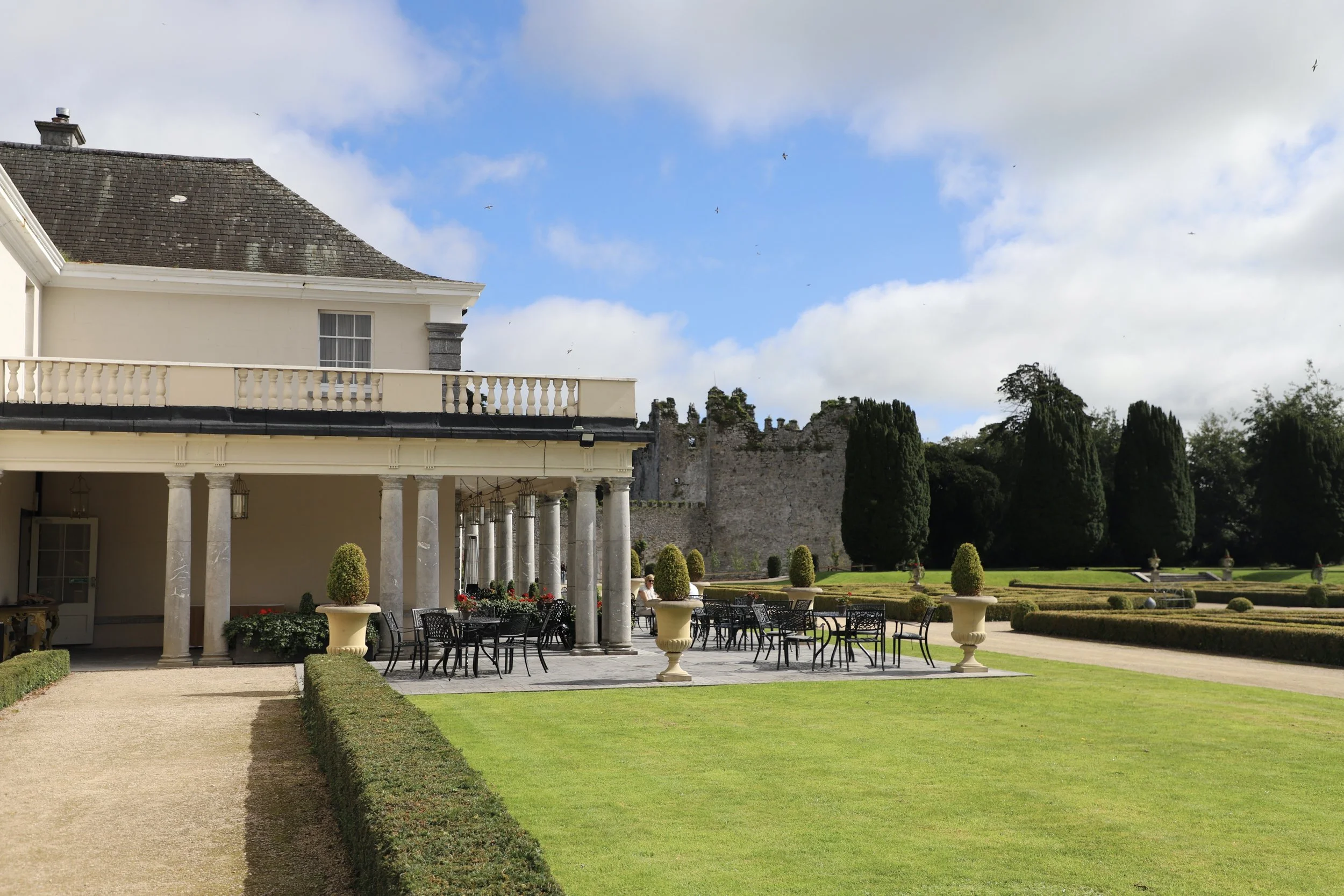 A spacious garden with a patio area featuring black metal tables and chairs, large potted plants, and a covered porch with classical columns. In the background, there is a stone castle wall surrounded by tall, dark green trees under a partly cloudy blue sky.
