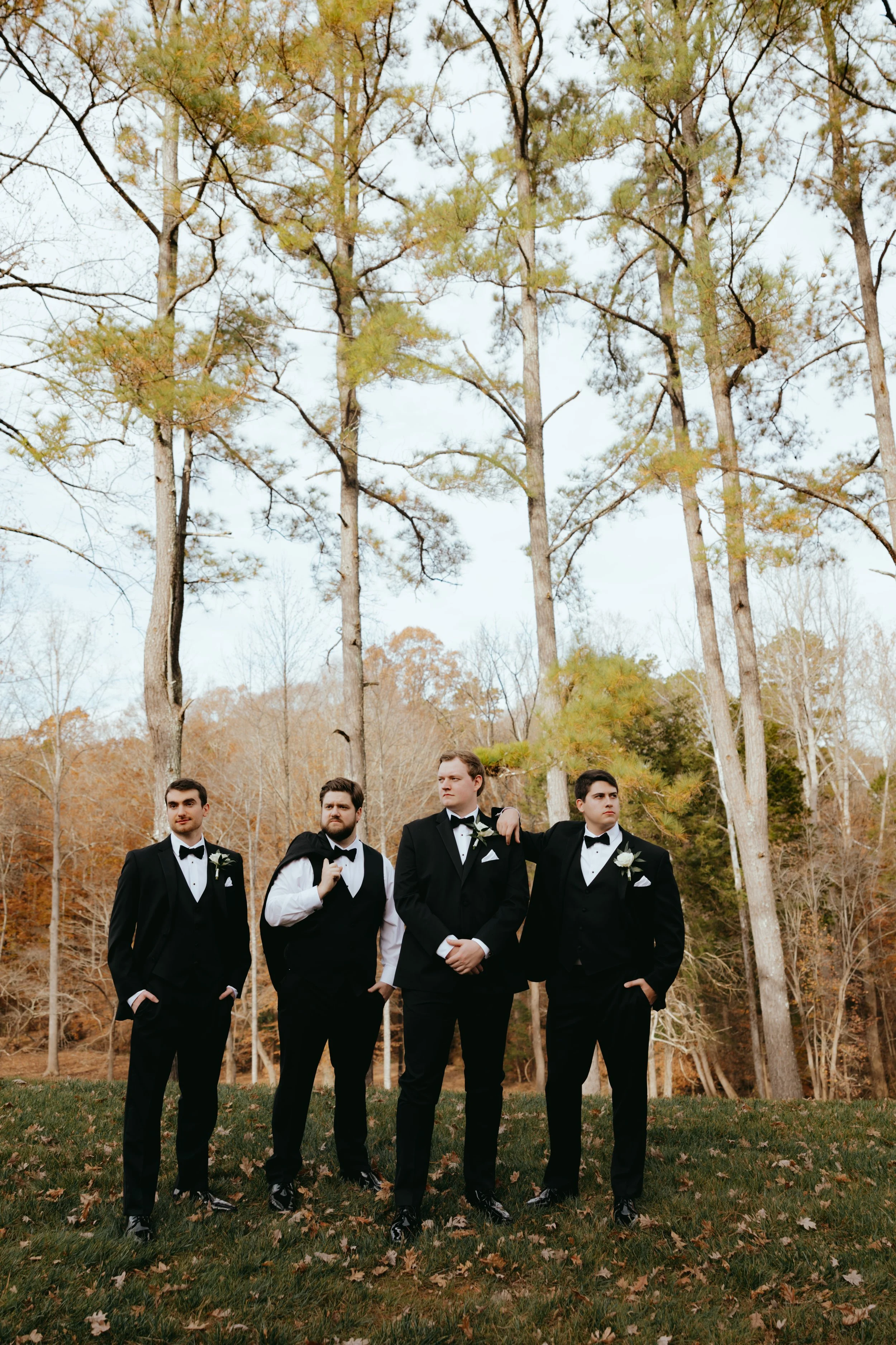 Four men in tuxedos standing outdoors on grass with autumn trees in the background.