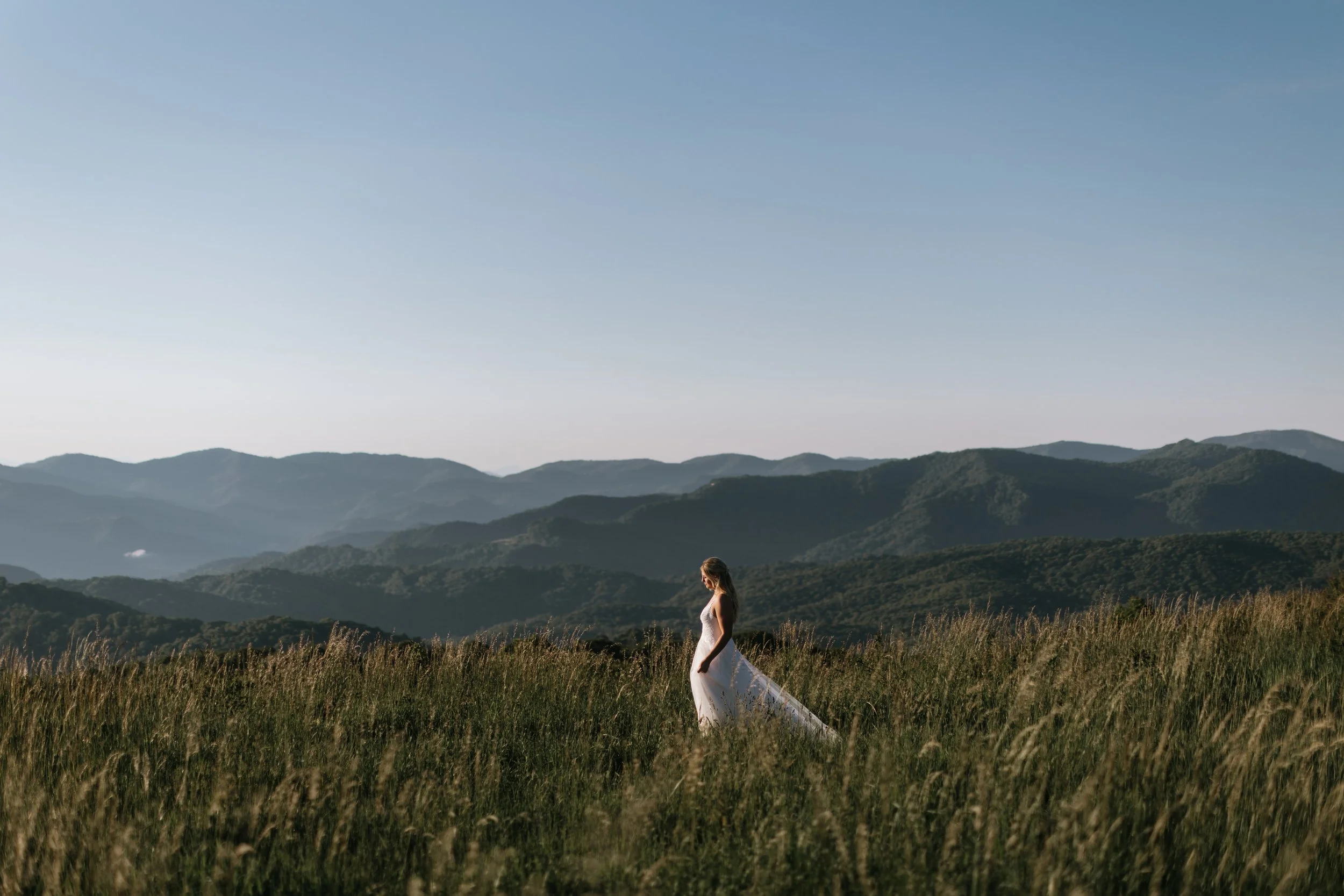Woman in a white dress standing in a grassy field with mountains in the background under a clear blue sky.