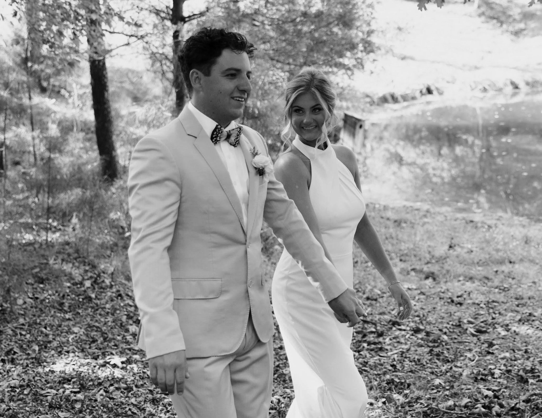 A black-and-white photo of a newlywed couple walking outdoors near a lake, smiling at each other. The groom is wearing a light-colored suit with a bowtie, and the bride is in a sleeveless white gown.