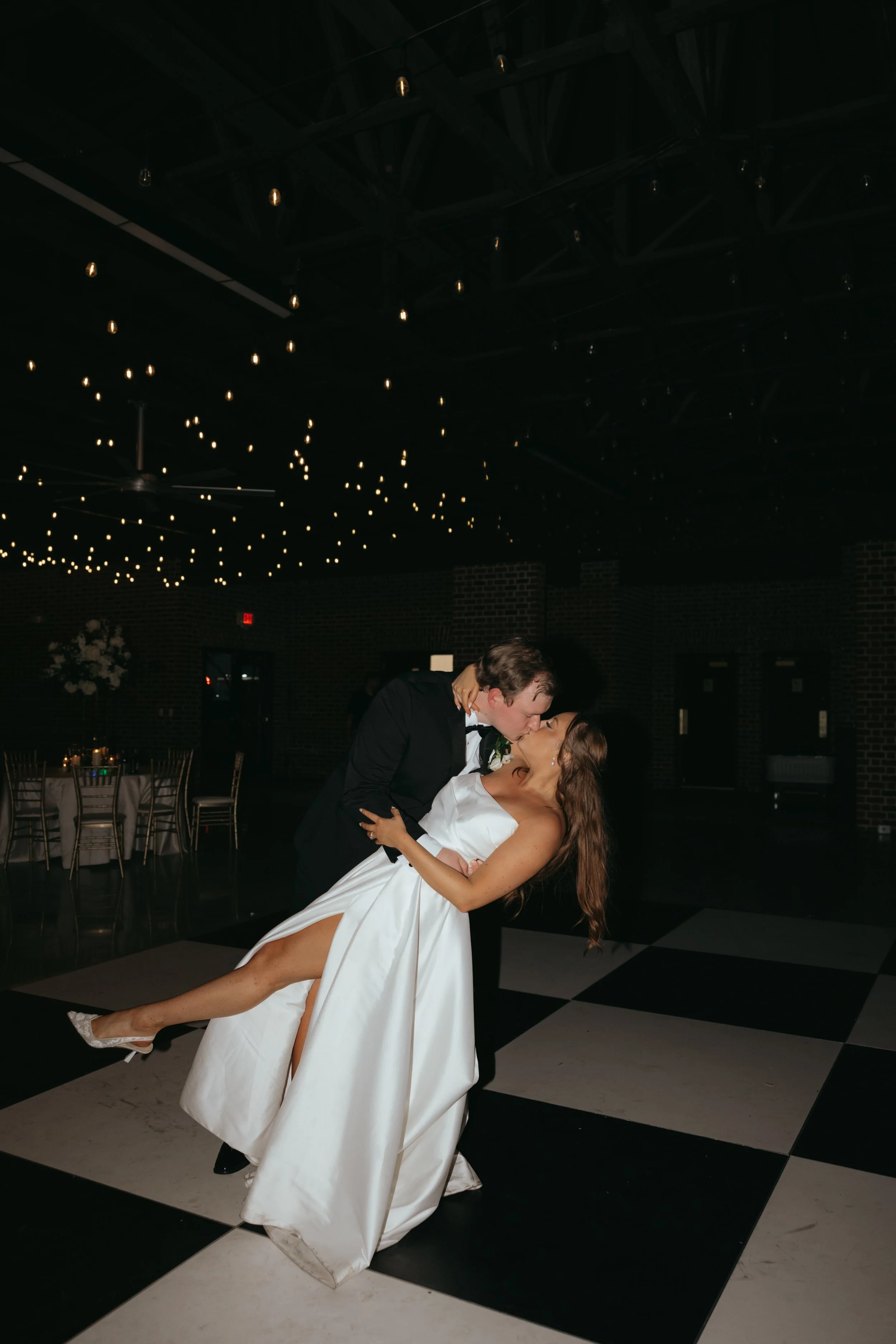 A newlywed couple sharing a kiss while dancing at their wedding reception, with the groom dipping the bride on a black-and-white checkered dance floor, decorated with a ceiling featuring string lights.