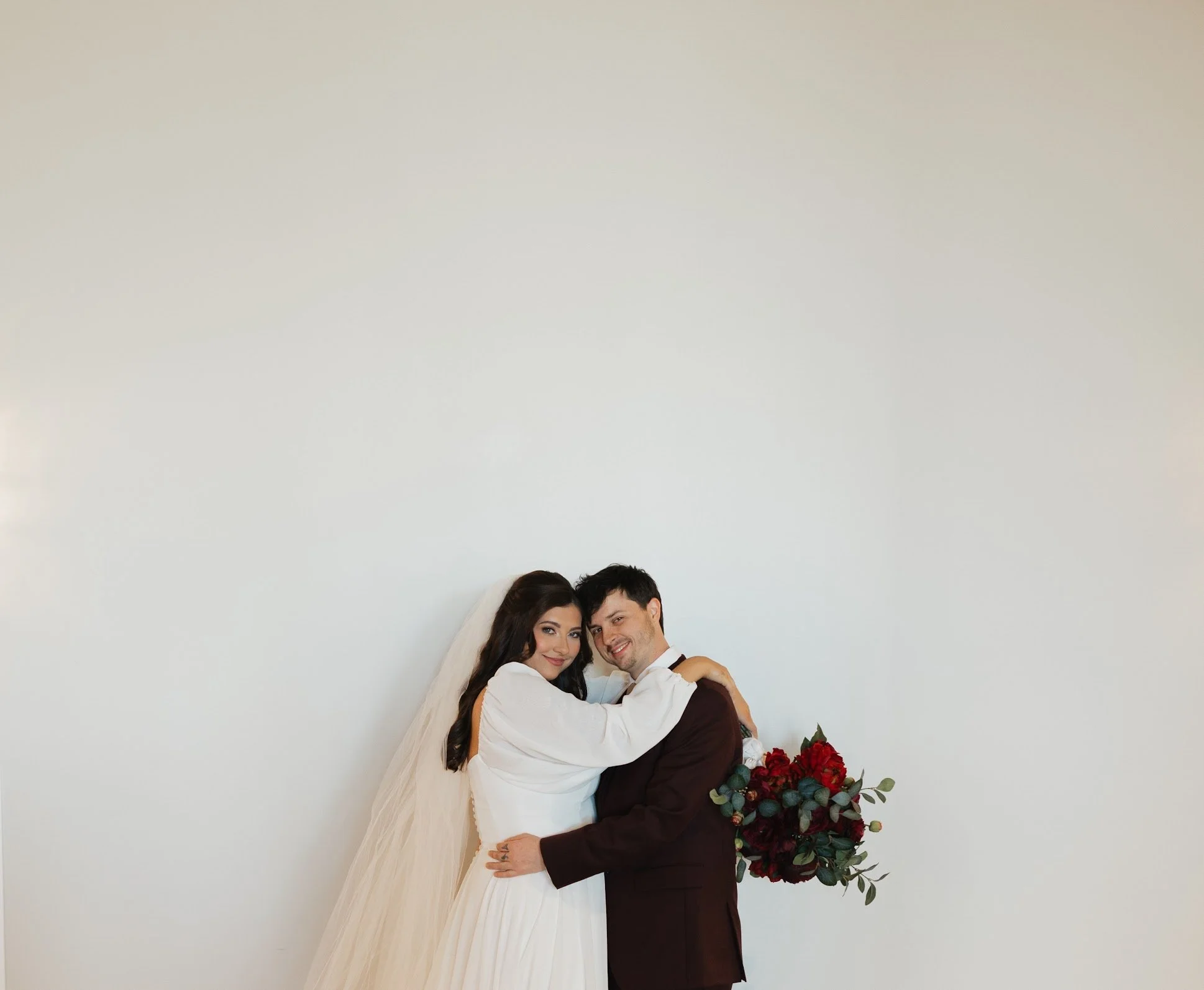 A bride and groom embracing for a photo, with the bride holding a bouquet of red roses and greenery, standing against a plain white background.