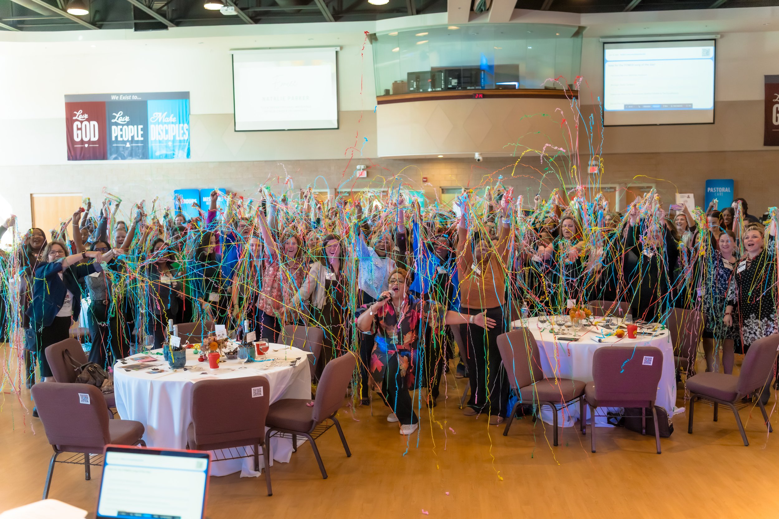 Large group of people celebrating at an event with colorful streamers, some seated at round tables and others standing, in an indoor venue with banners on the wall.