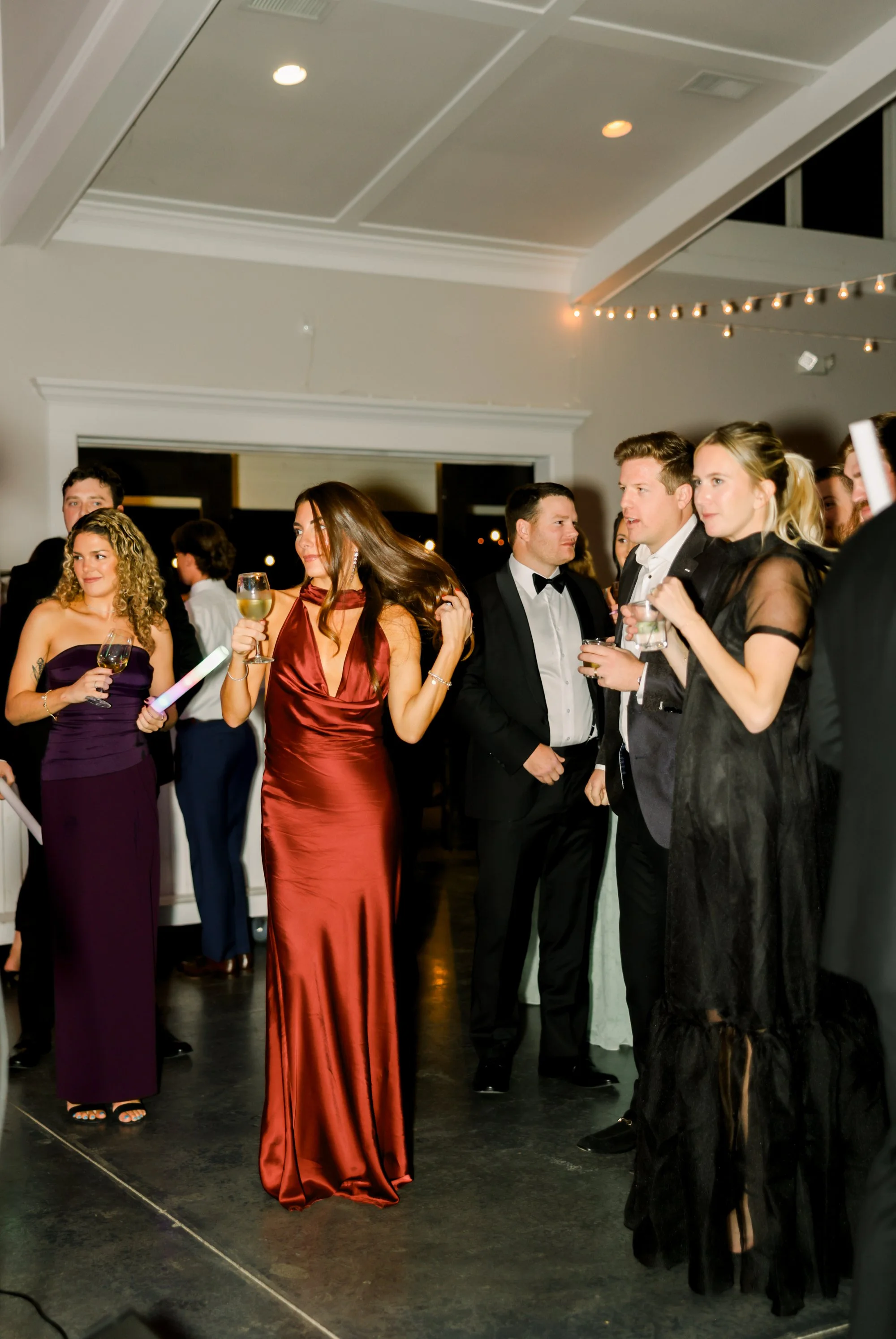 People dressed in formal evening wear at a party, holding drinks, in a decorated indoor space with string lights.