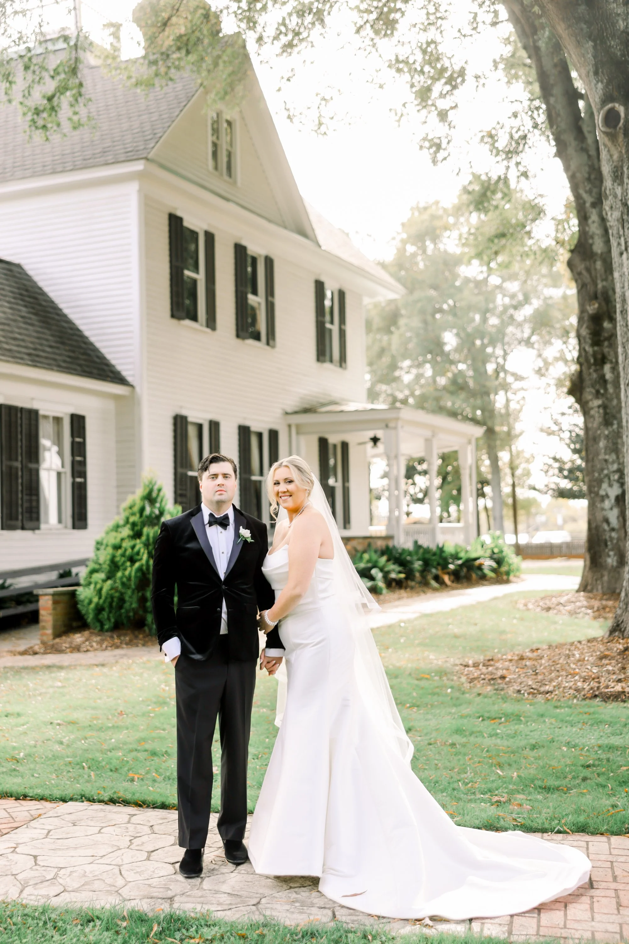 Bride and groom standing outside in wedding attire in front of a large white house with black shutters, trees, and greenery.
