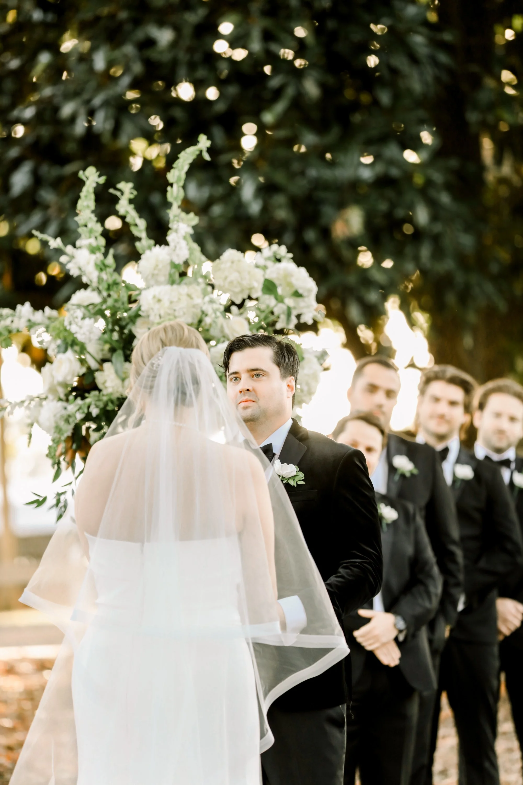 Bride and groom exchanging vows during outdoor wedding ceremony, with groomsmen standing in the background and floral arrangements behind them.