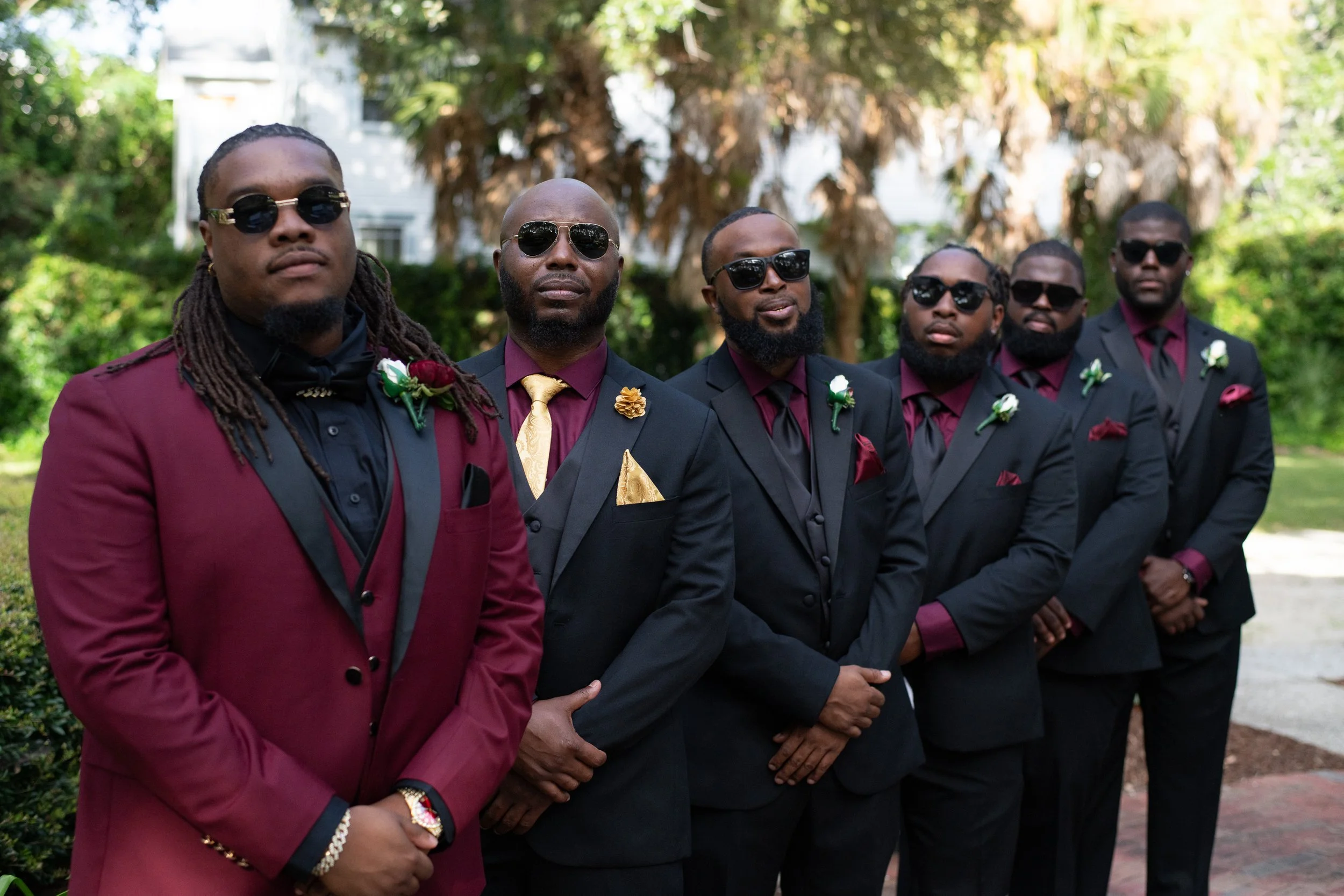A group of six men dressed in formal suits standing outdoors, all wearing dark sunglasses and boutonnières or lapel flowers, with greenery and trees in the background.