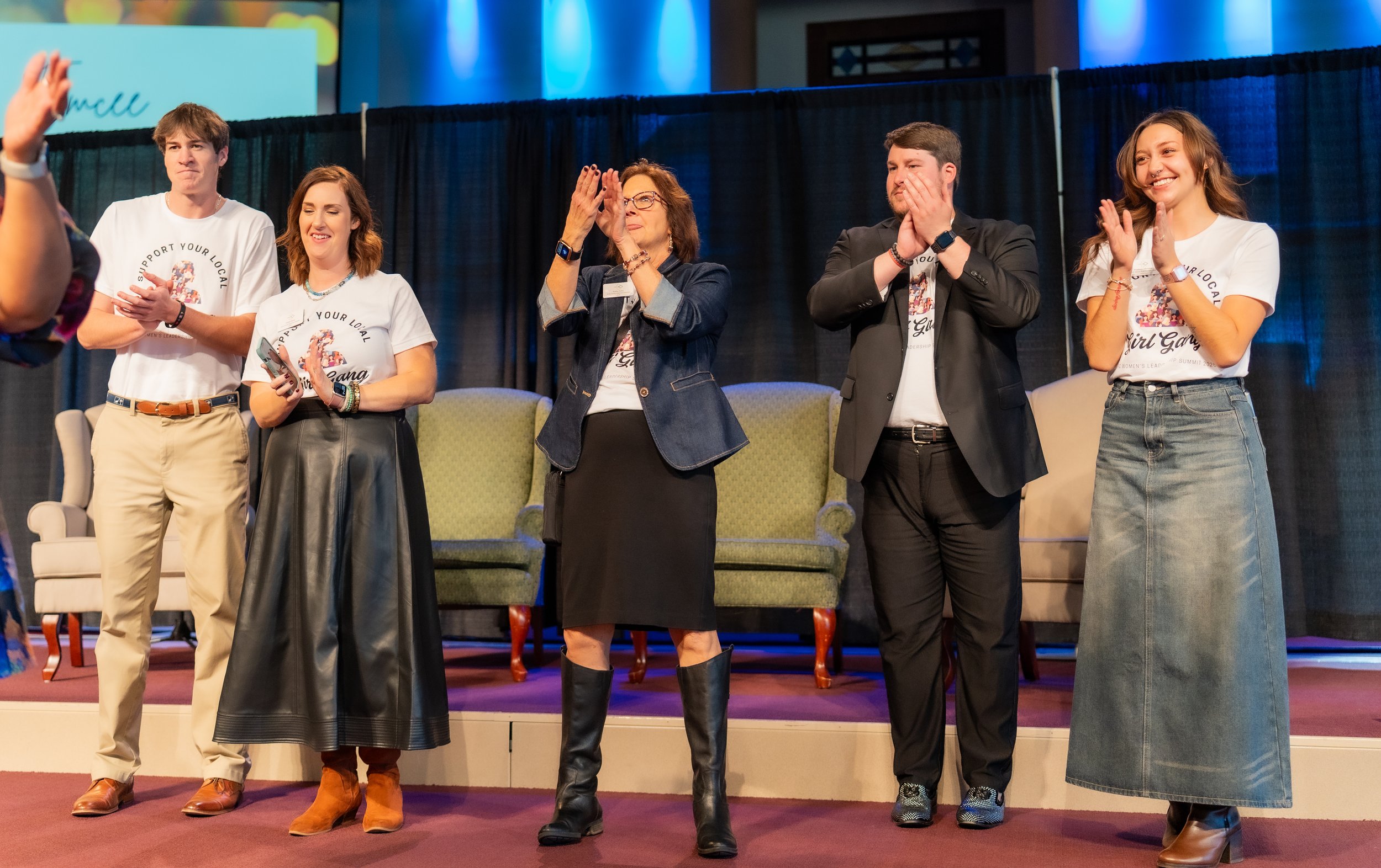 Group of five people standing on a stage, applauding, with black curtains and a green sofa behind them, during an event or conference.