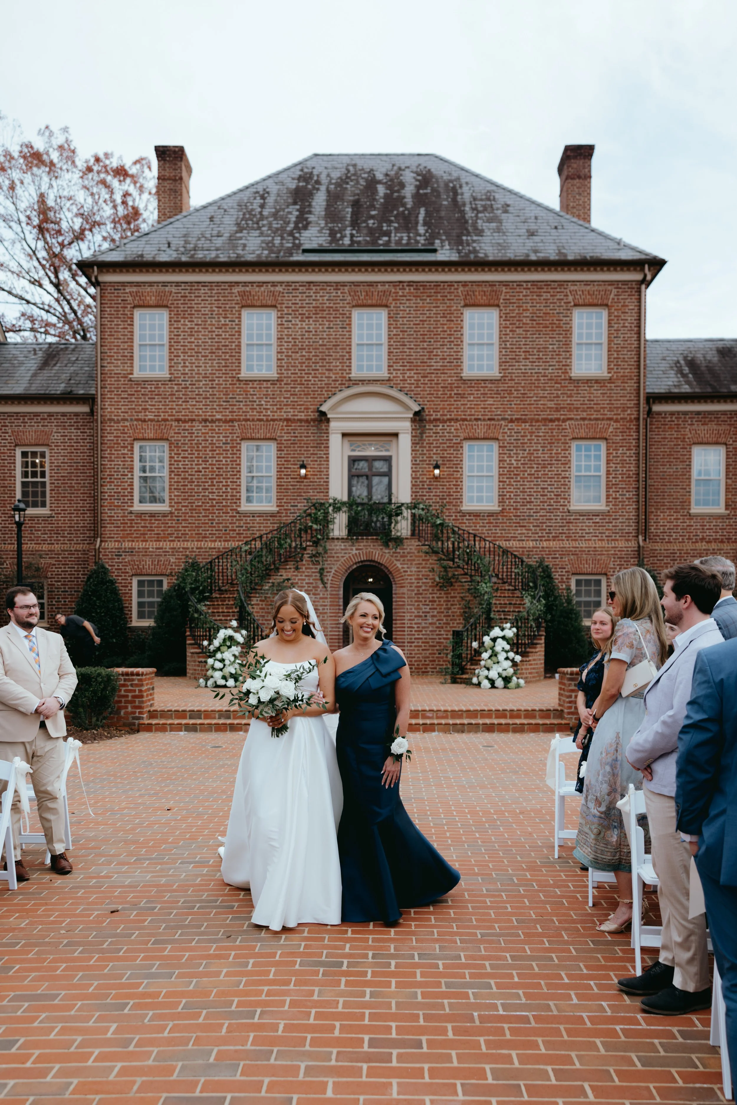A wedding ceremony taking place outdoors in front of a large brick mansion. Brides are walking down the aisle, with one in a white wedding dress holding a bouquet, and the other in a blue gown. Guests are standing on either side, watching and smiling