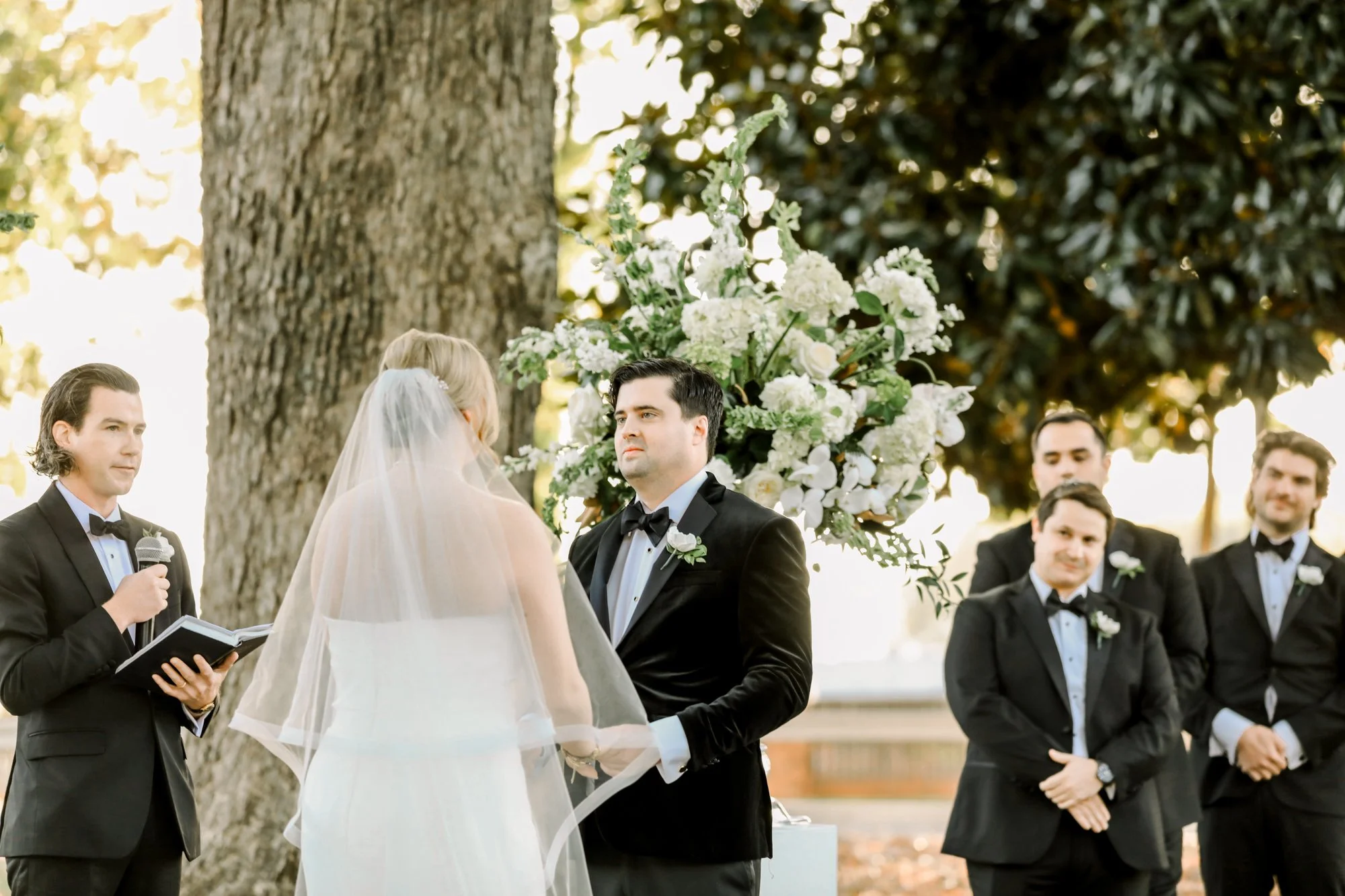 A wedding ceremony outdoors with a bride and groom exchanging vows. The bride wears a white dress and veil, and the groom wears a black tuxedo. An officiant holding a microphone and book stands nearby. Four groomsmen in tuxedos stand in the backgroun