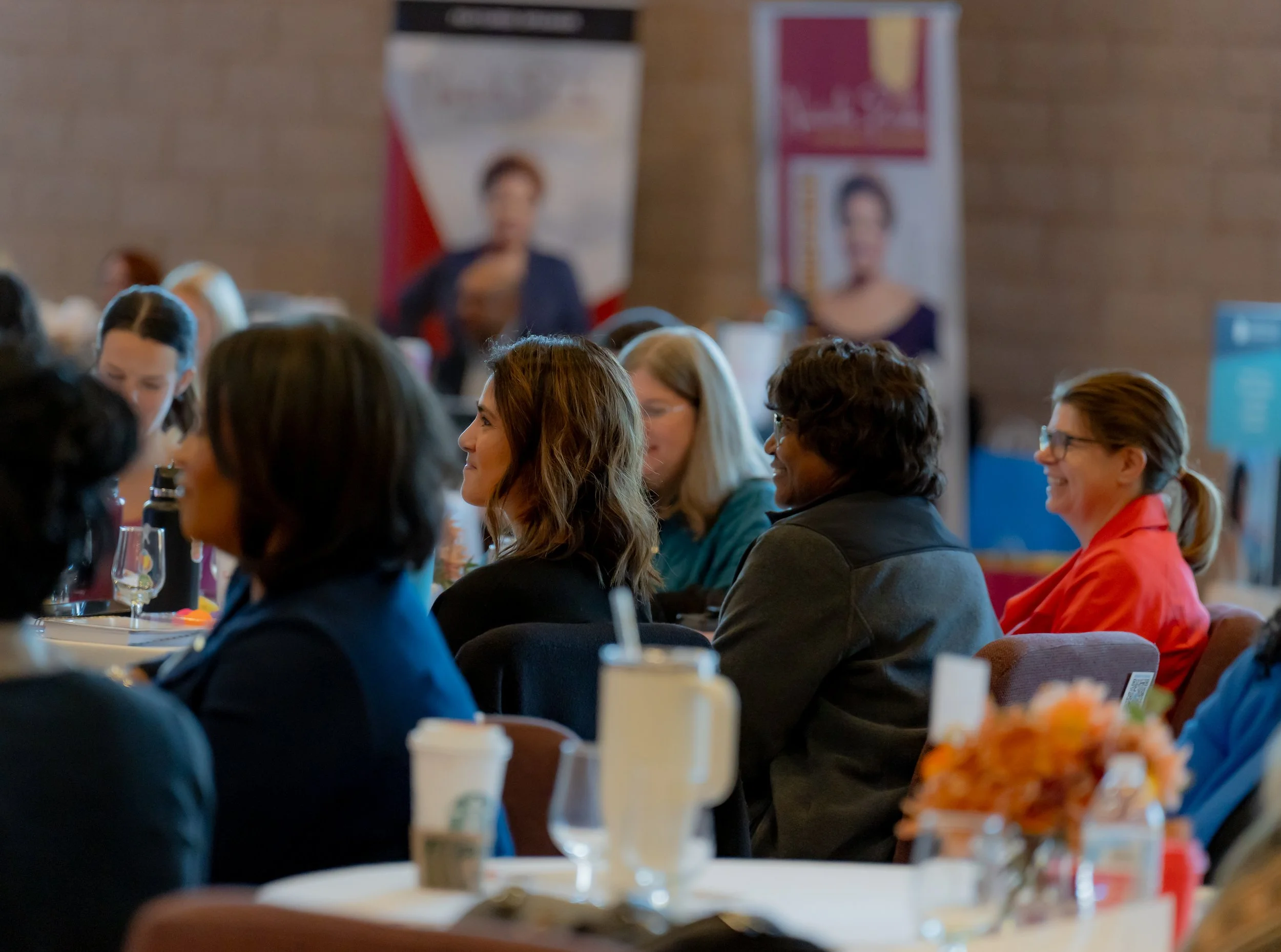A group of women seated at a conference table, engaged and smiling, in a professional setting with banners and informational displays in the background.