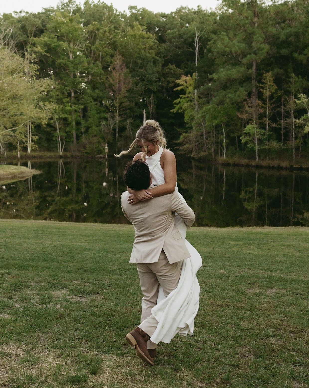 A man lifting a woman in a white dress by a lake with trees in the background.