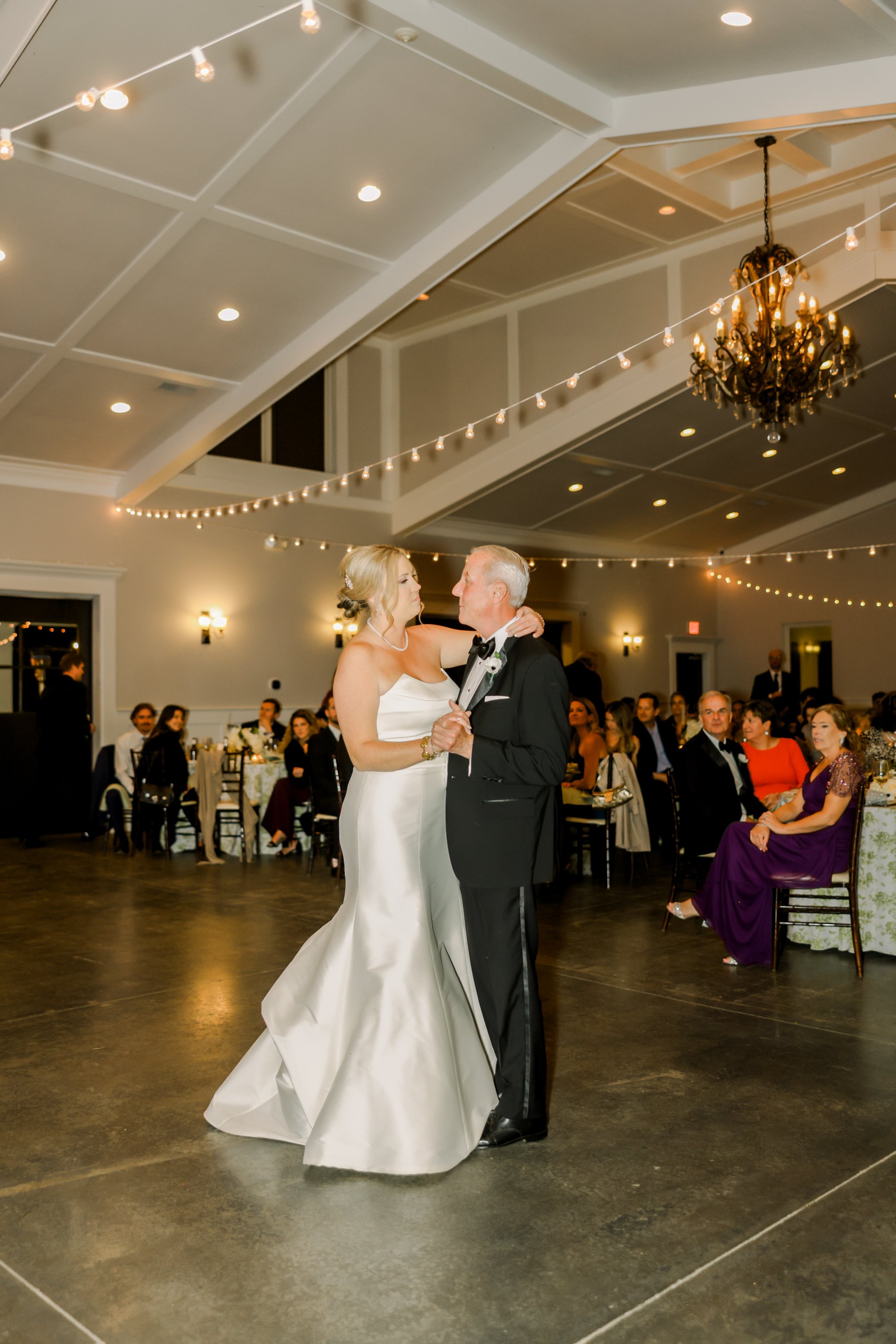 A bride and an older man, possibly her father, sharing a dance at a wedding reception in a decorated banquet hall with guests watching.