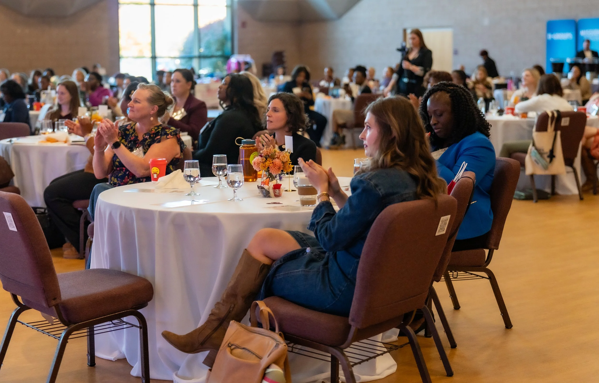 A large group of women attending a conference or seminar, sitting around round tables with white tablecloths, listening and clapping, with some having drinks and snacks, in a well-lit room with large windows and a stage or presentation area at the fr