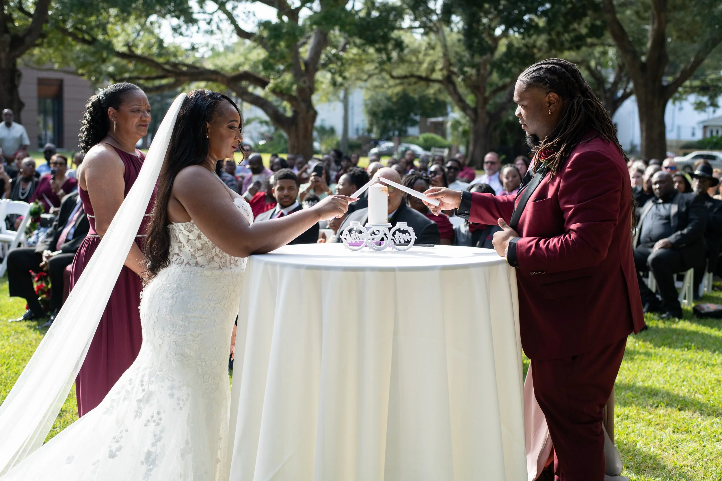 A couple exchanging wedding vows outdoors under a tree, with guests seated behind them. The bride wears a white lace gown and veil, while the groom wears a burgundy suit. They stand at a round table with a candle and decorative elements.