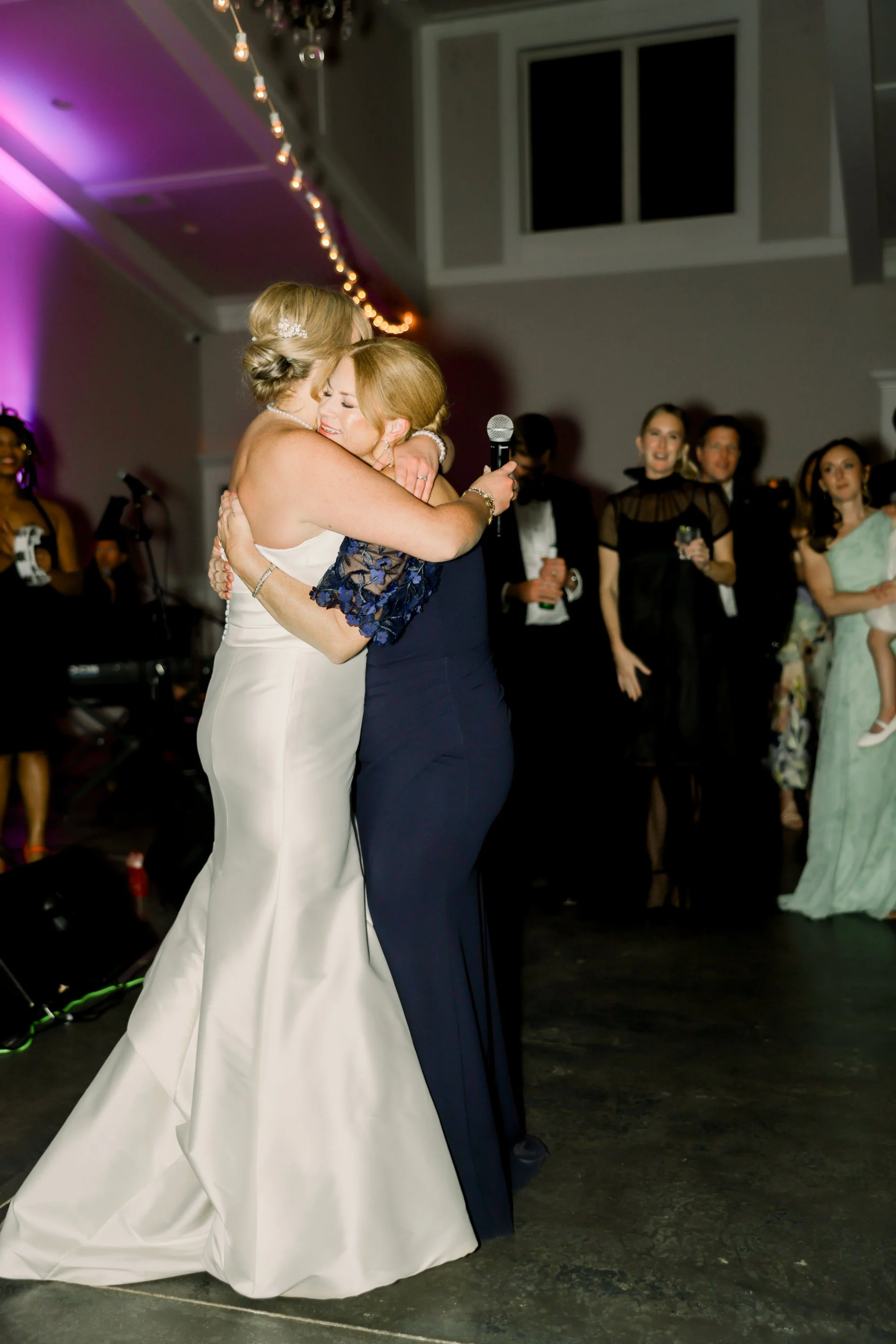 Two women are hugging at a wedding celebration. One is dressed in a white wedding gown, and the other in a dark blue dress. They are on a dance floor with guests and a band in the background.