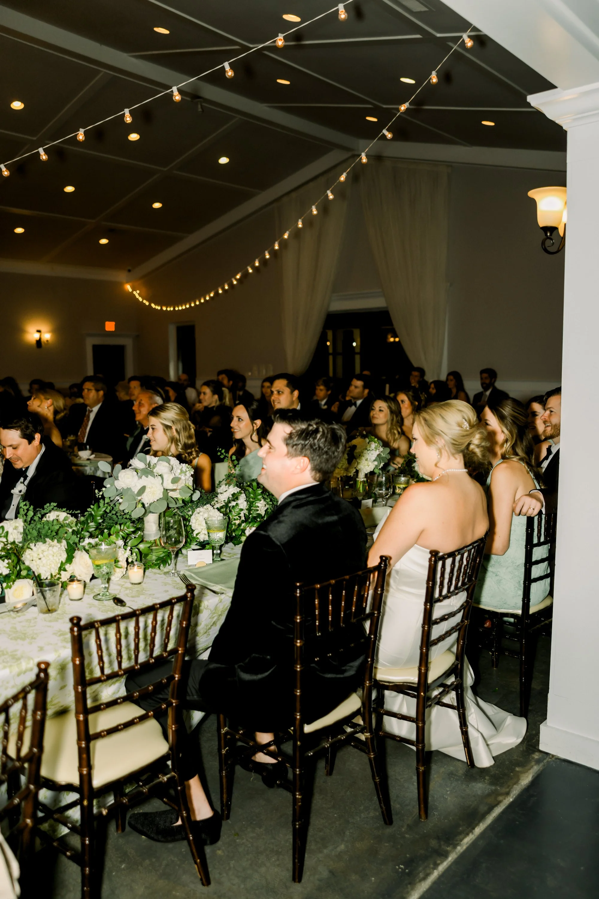 Guests seated at a wedding reception table, decorated with white flowers and candles, under hanging string lights in a dimly lit room.