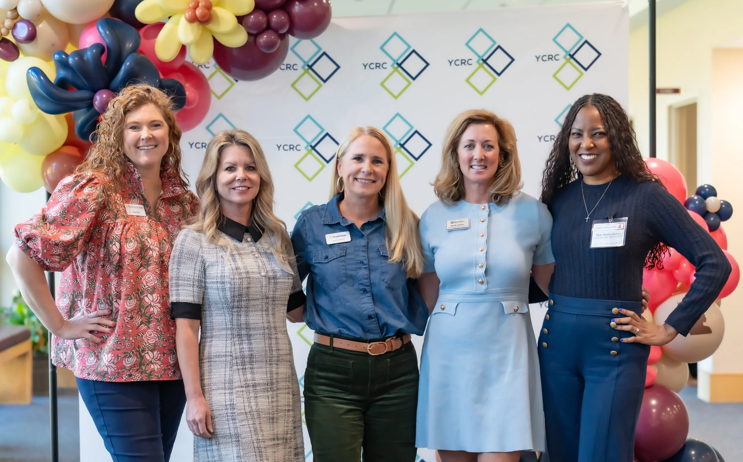 Five women standing side by side in front of a backdrop with a colorful balloon arch to the left and the YCRC logo. They are dressed professionally and smiling at the camera.