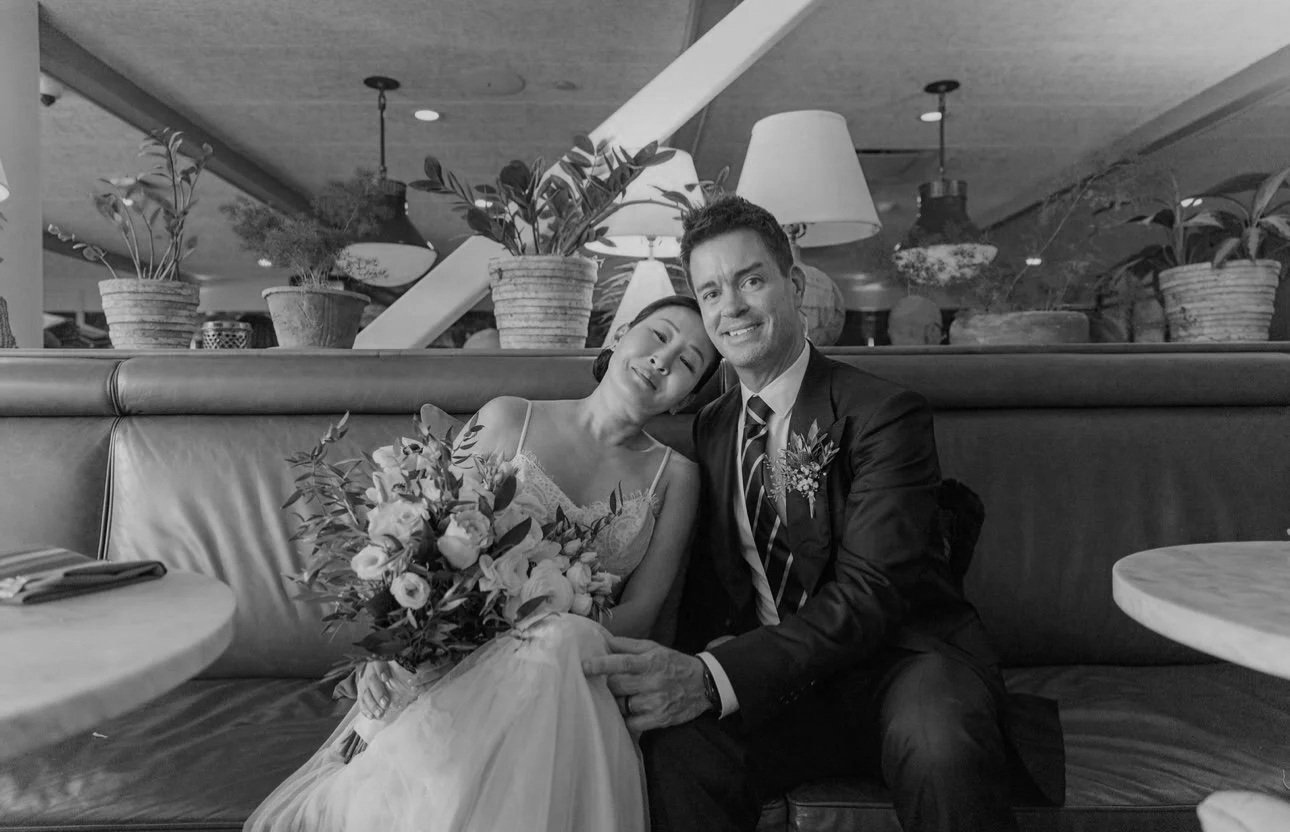 A bride and groom sitting on a booth in a restaurant, smiling, with the bride holding a bouquet and leaning her head on the groom's shoulder.