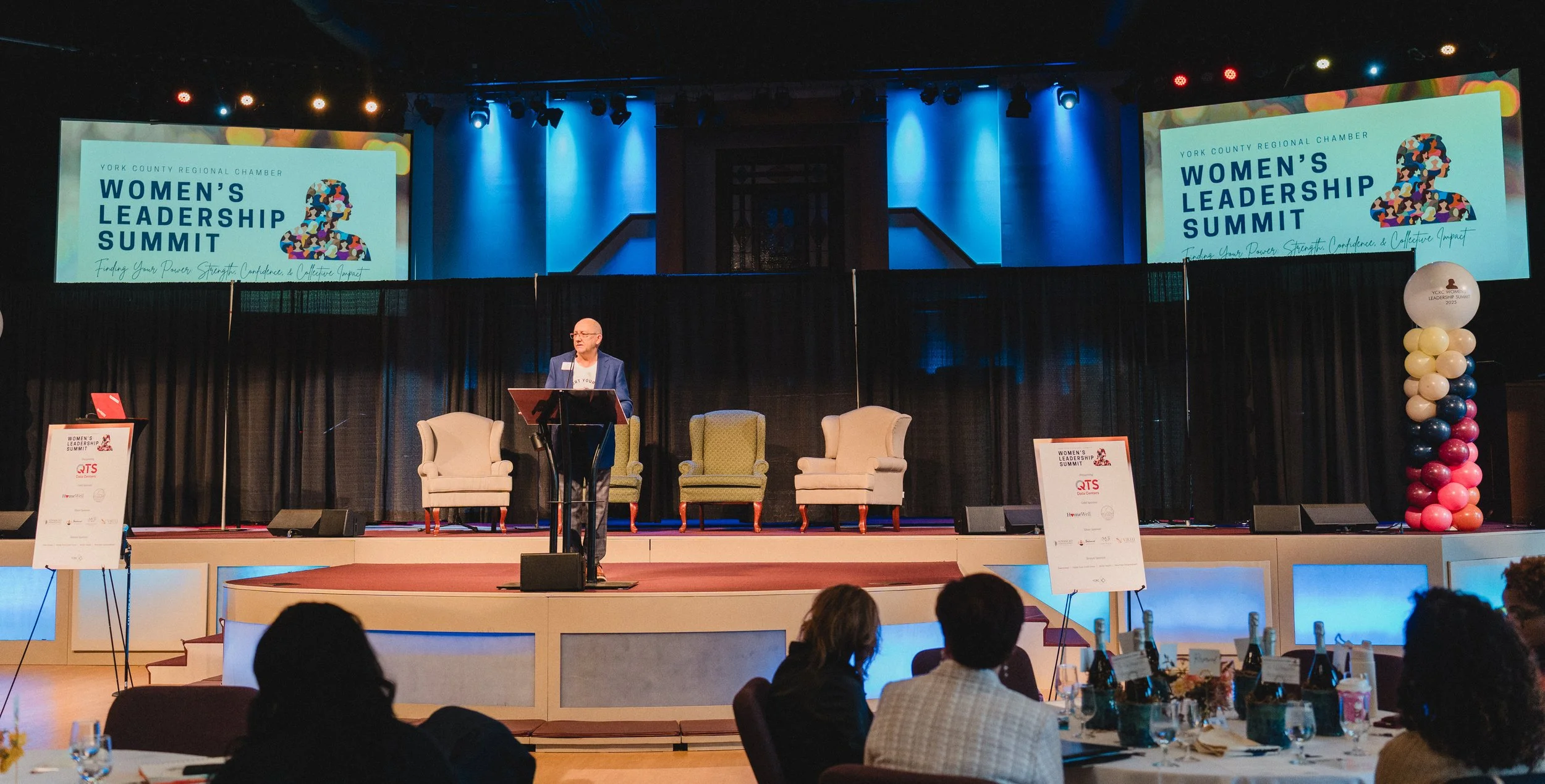 A speaker at a podium addresses attendees during the Women's Leadership Summit in a large conference hall. The stage features four armchairs, and large screens display the event's logo and theme. The room has colorful balloon decorations.