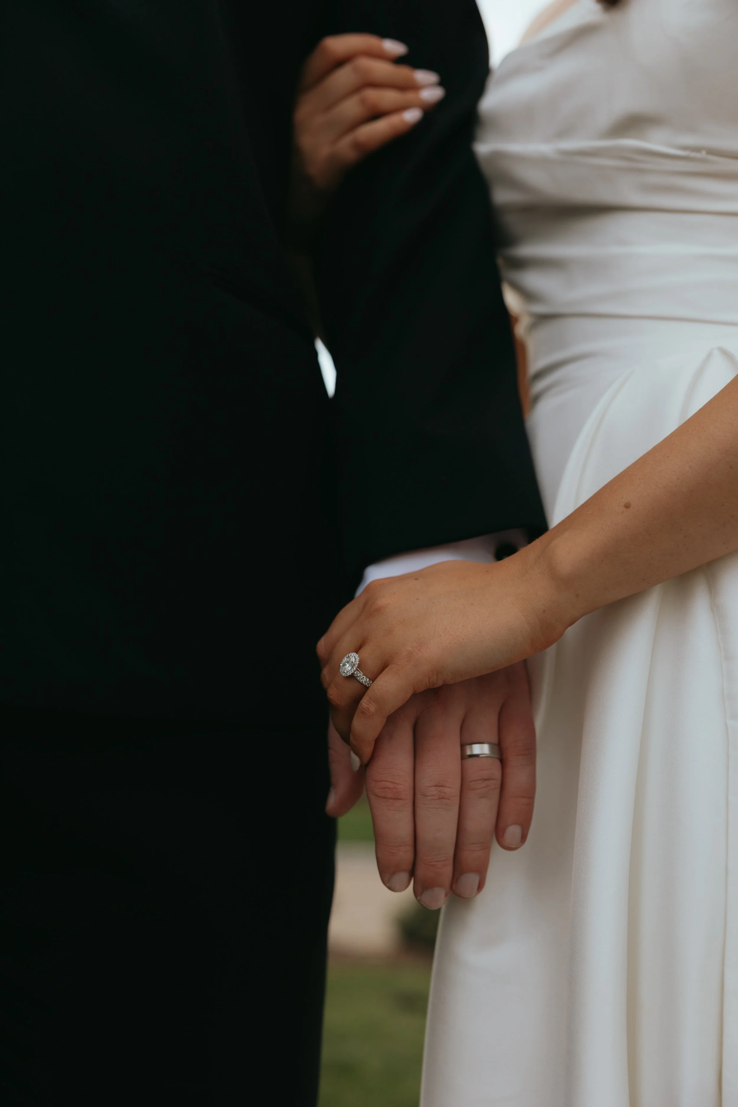 Close-up of a couple holding hands, with wedding rings visible on their ring fingers. The person on the right is wearing a white dress and the person on the left wearing a black suit.