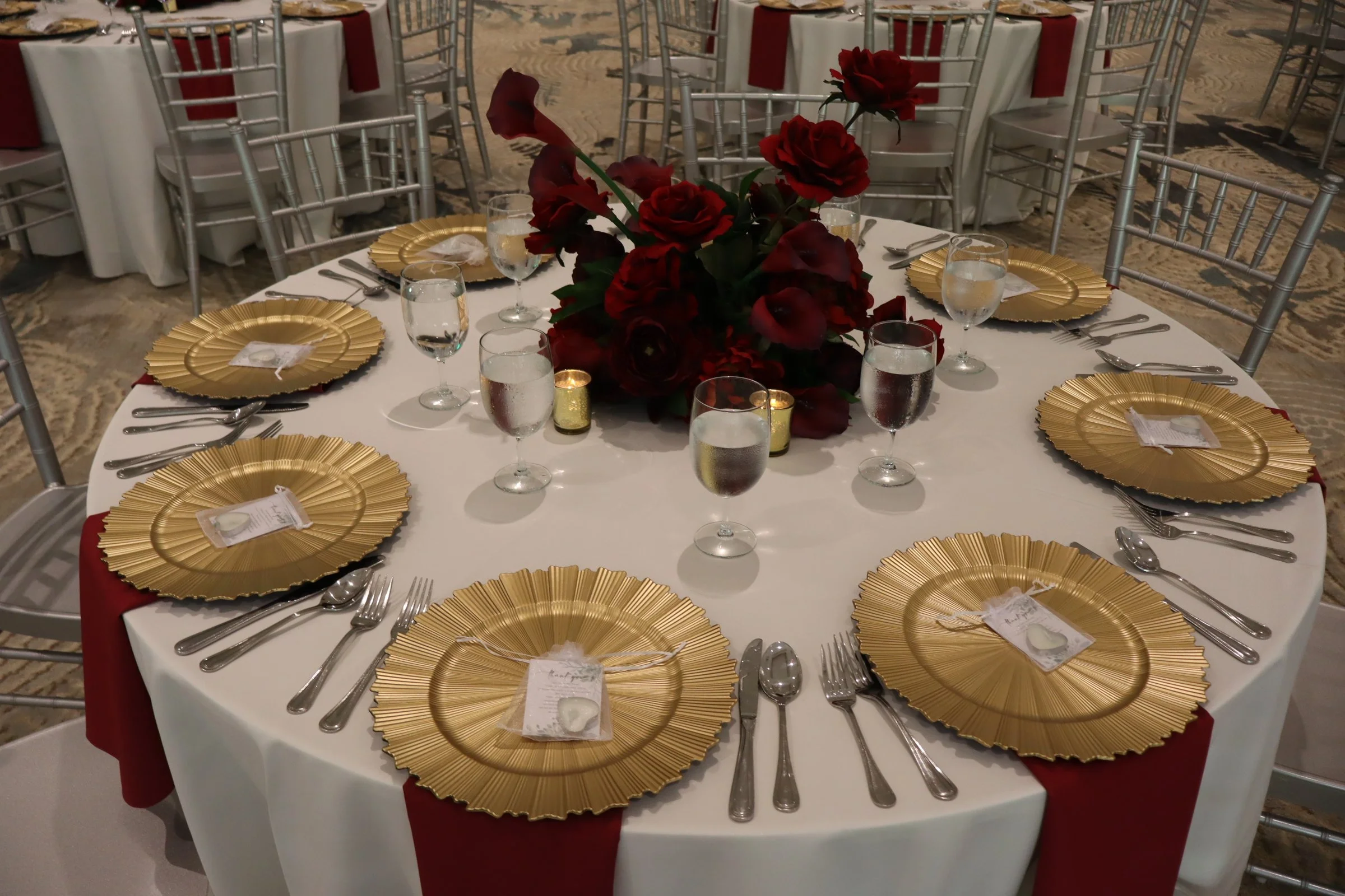 Round banquet table set for a formal event with gold-rimmed plates, water glasses, silverware, red napkins, and a centerpiece of dark red flowers. Gold candles are also on the table.