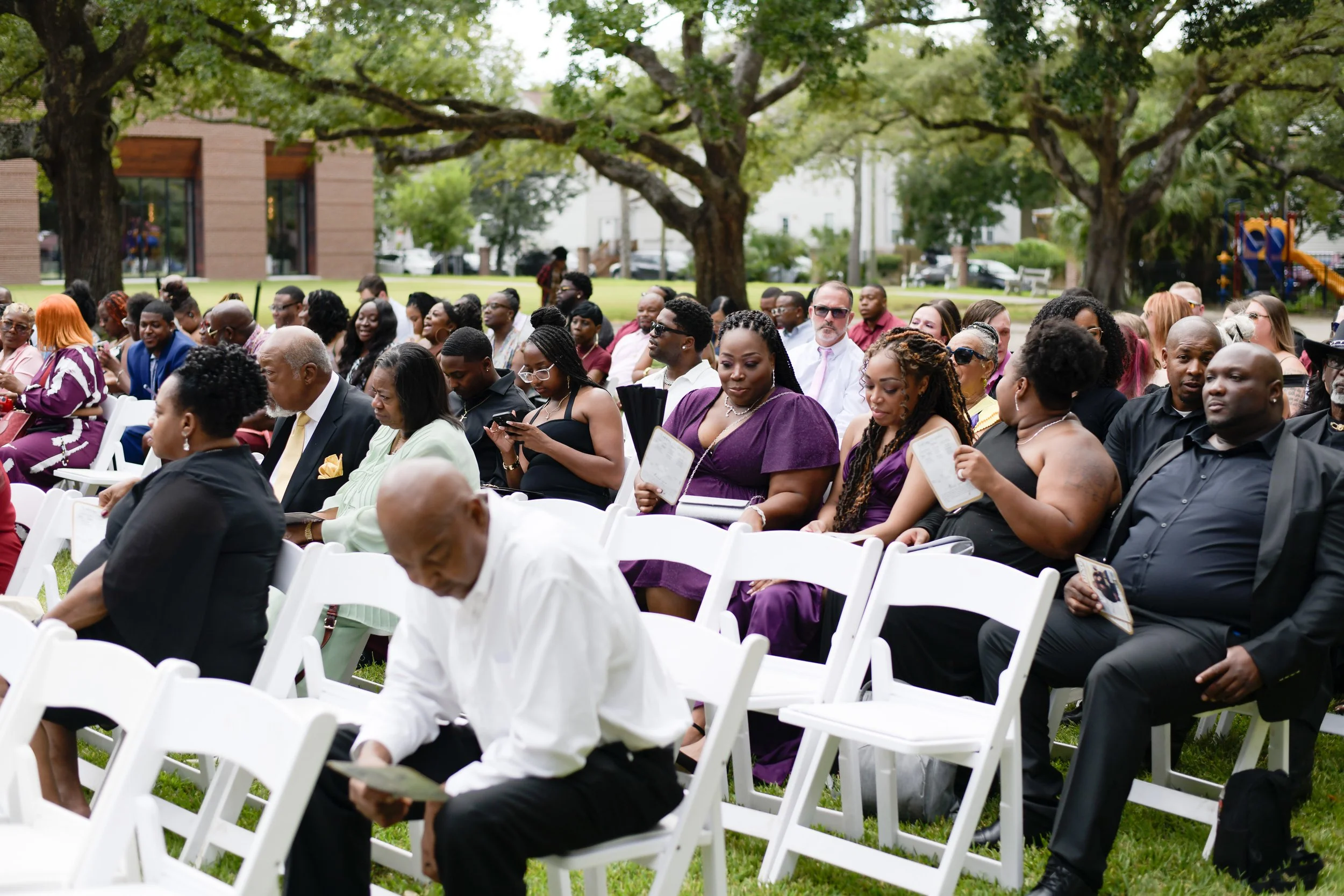 Group of diverse people seated outdoors at a formal event, dressed in elegant attire, with trees and a park in the background.