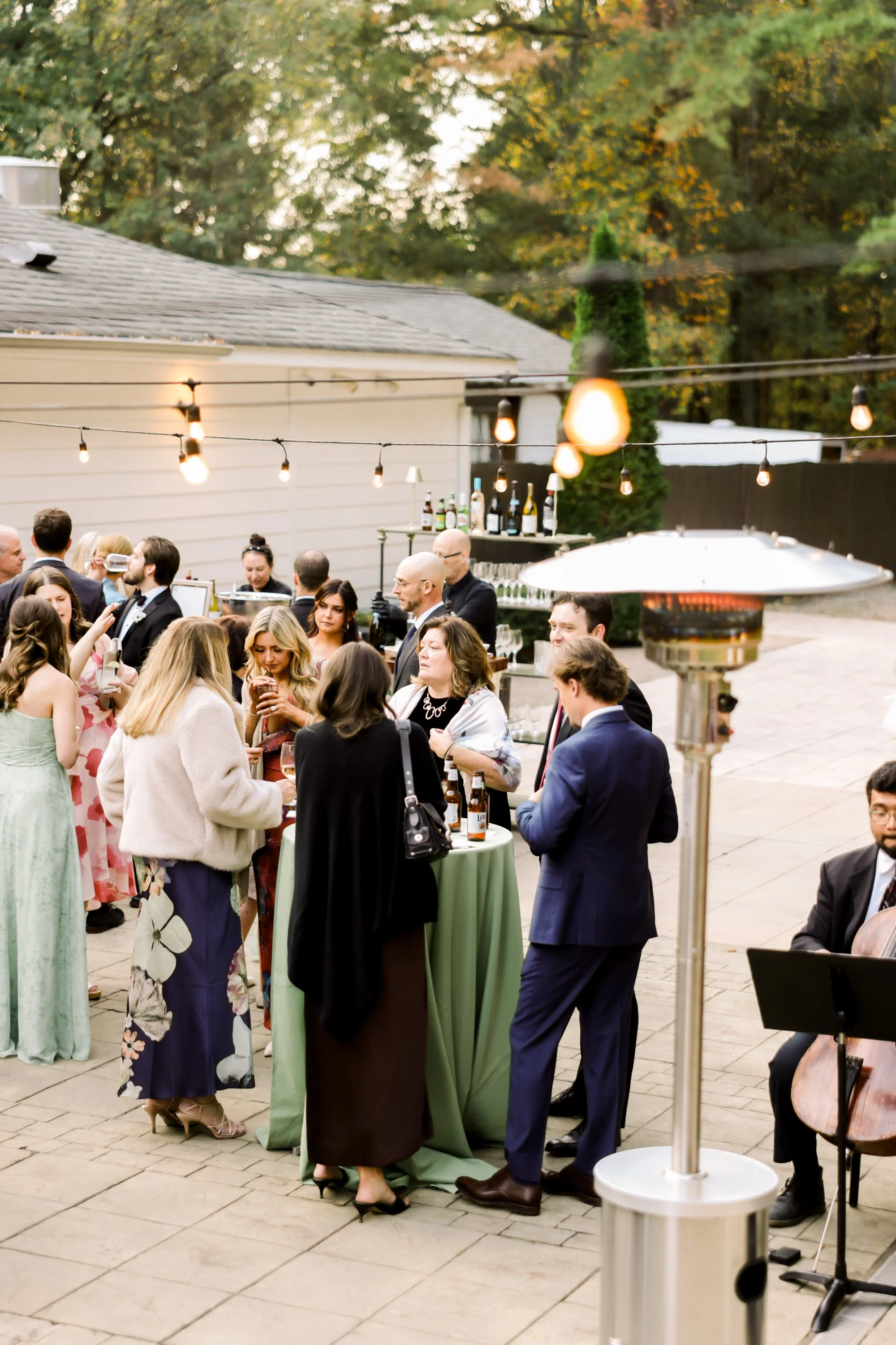 People at an outdoor gathering with string lights, a high-top table, a bar setup, a patio heater, and a musician playing a cello woman in the background.