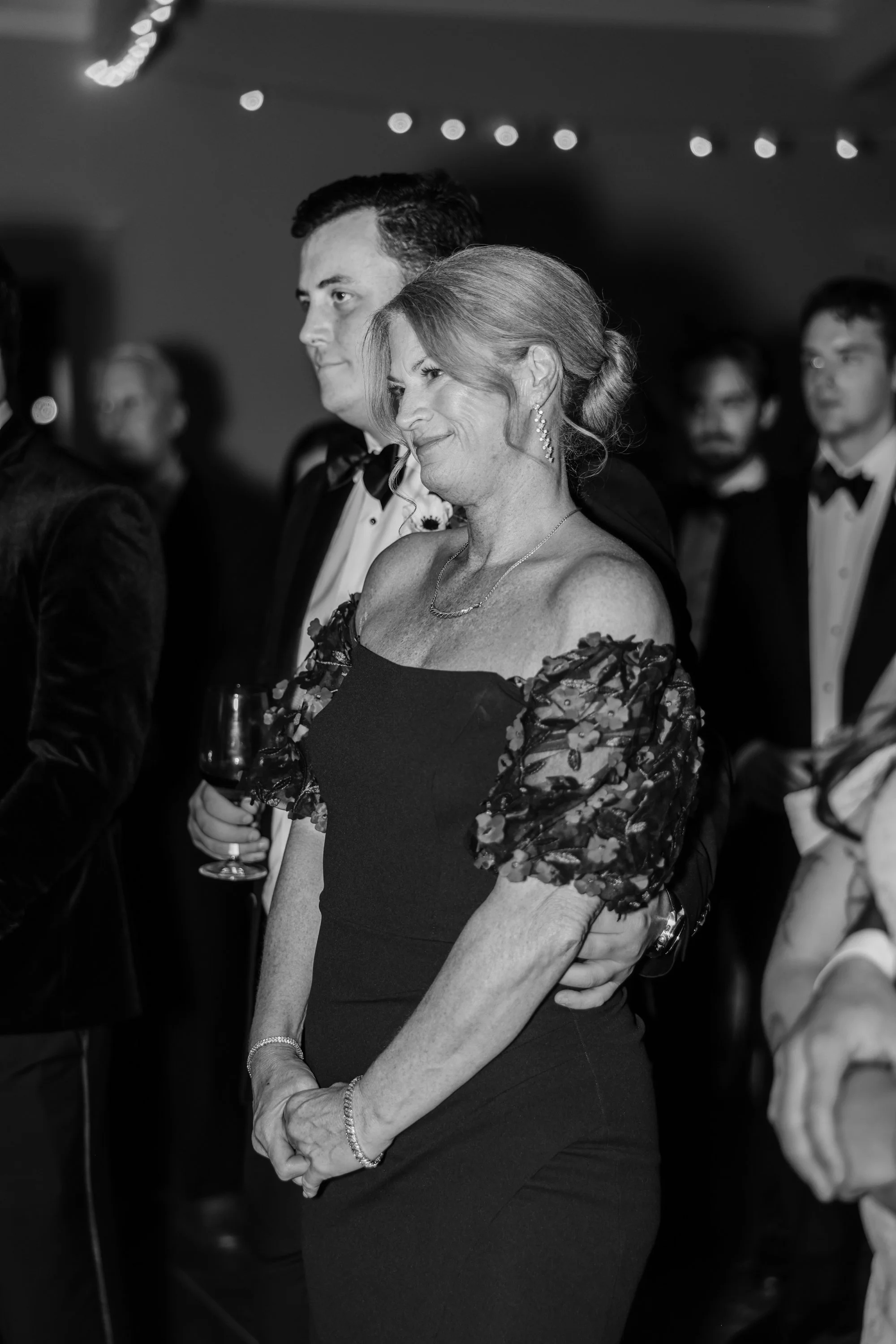 A woman in a black dress with floral sleeves stands among men in tuxedos at a formal event, holding a glass of wine and smiling lightly.