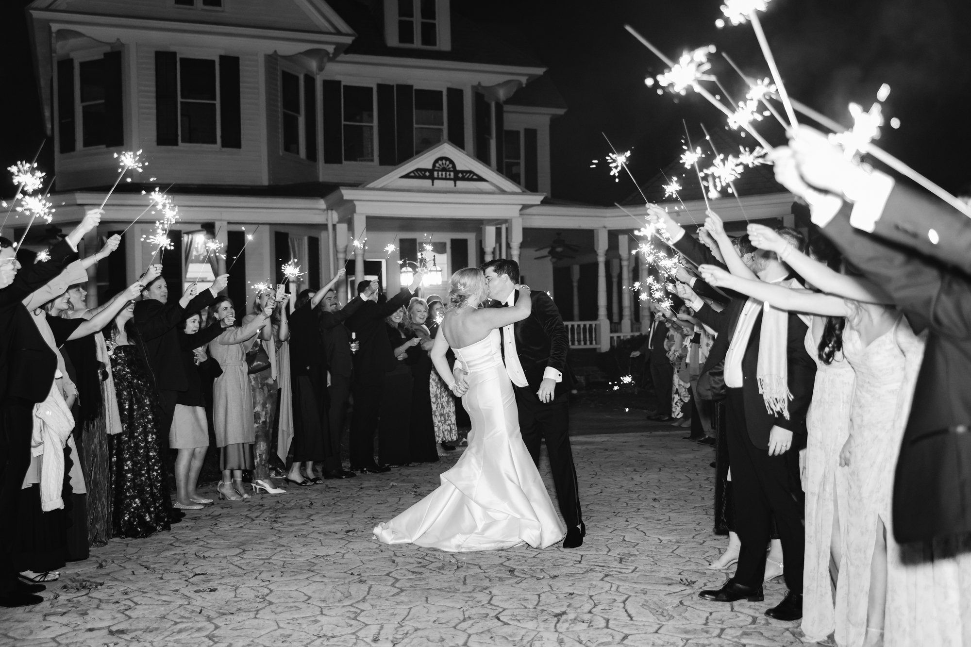 Black and white photo of a wedding reception with the bride and groom dancing in the center, surrounded by guests holding sparklers in front of a large house at night.