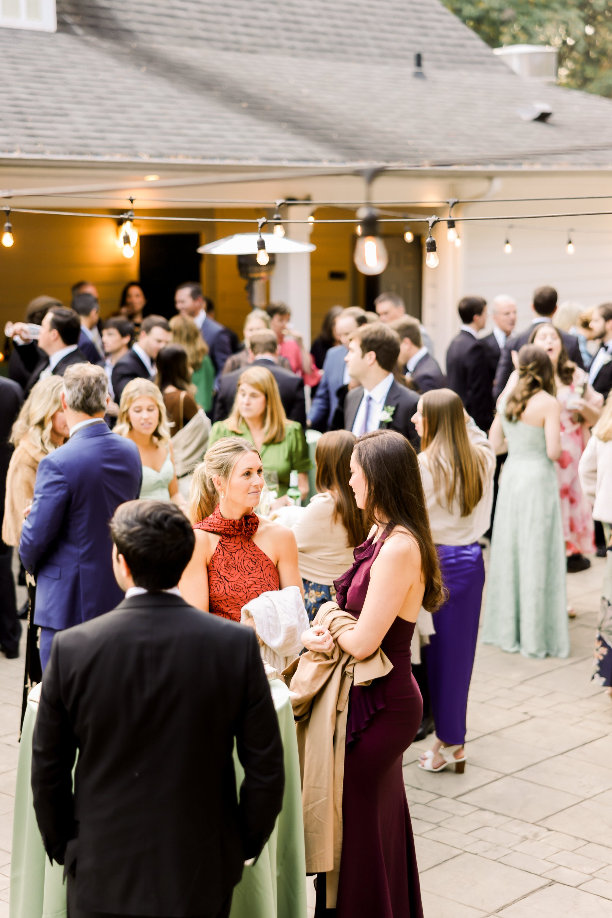 People in formal attire socializing outdoors at a gathering with string lights overhead.