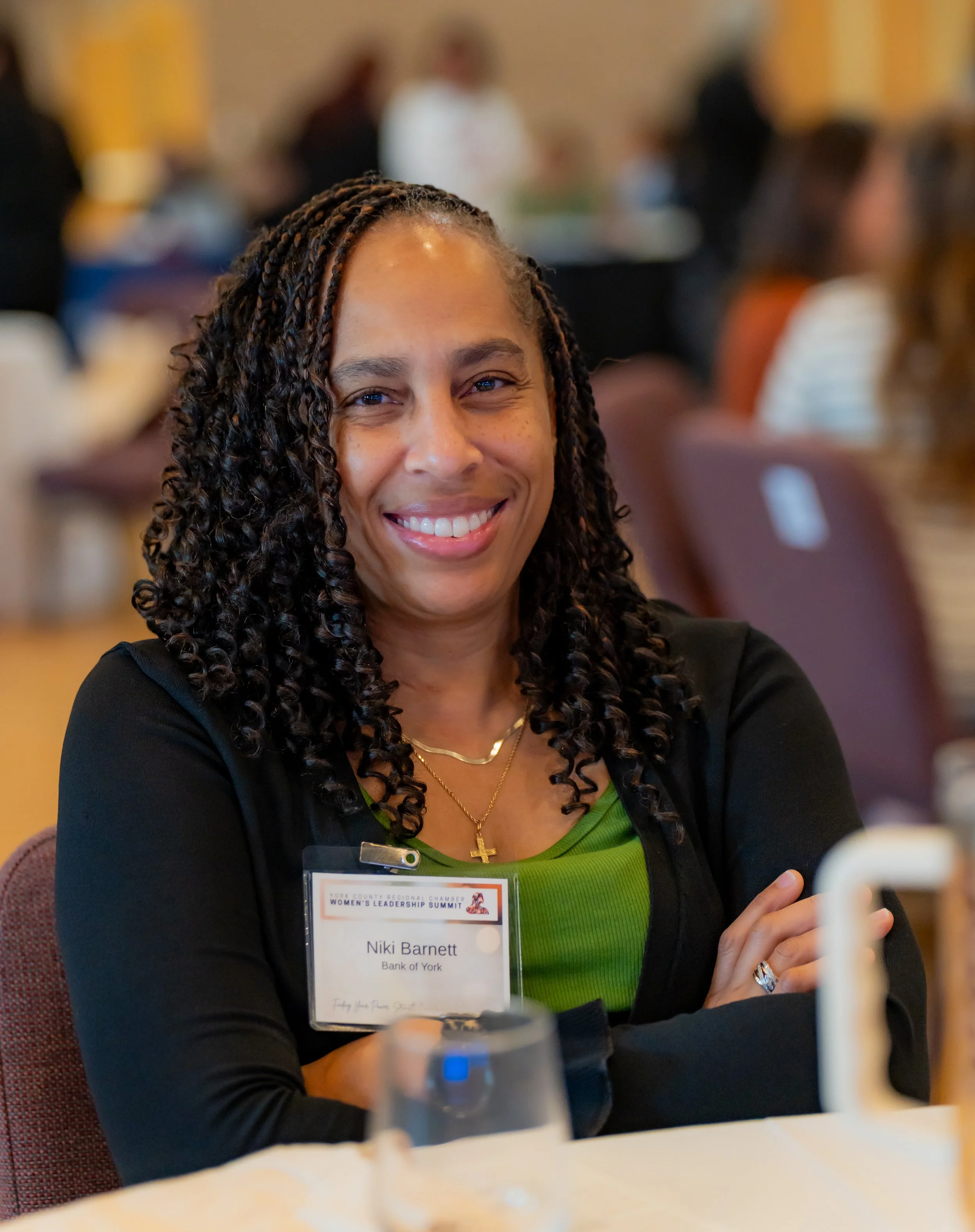 A woman with curly hair smiling at a conference, wearing a name badge that reads 'Niki Barnett, Bank of York,' dressed in a black blazer with a green top and gold jewelry.