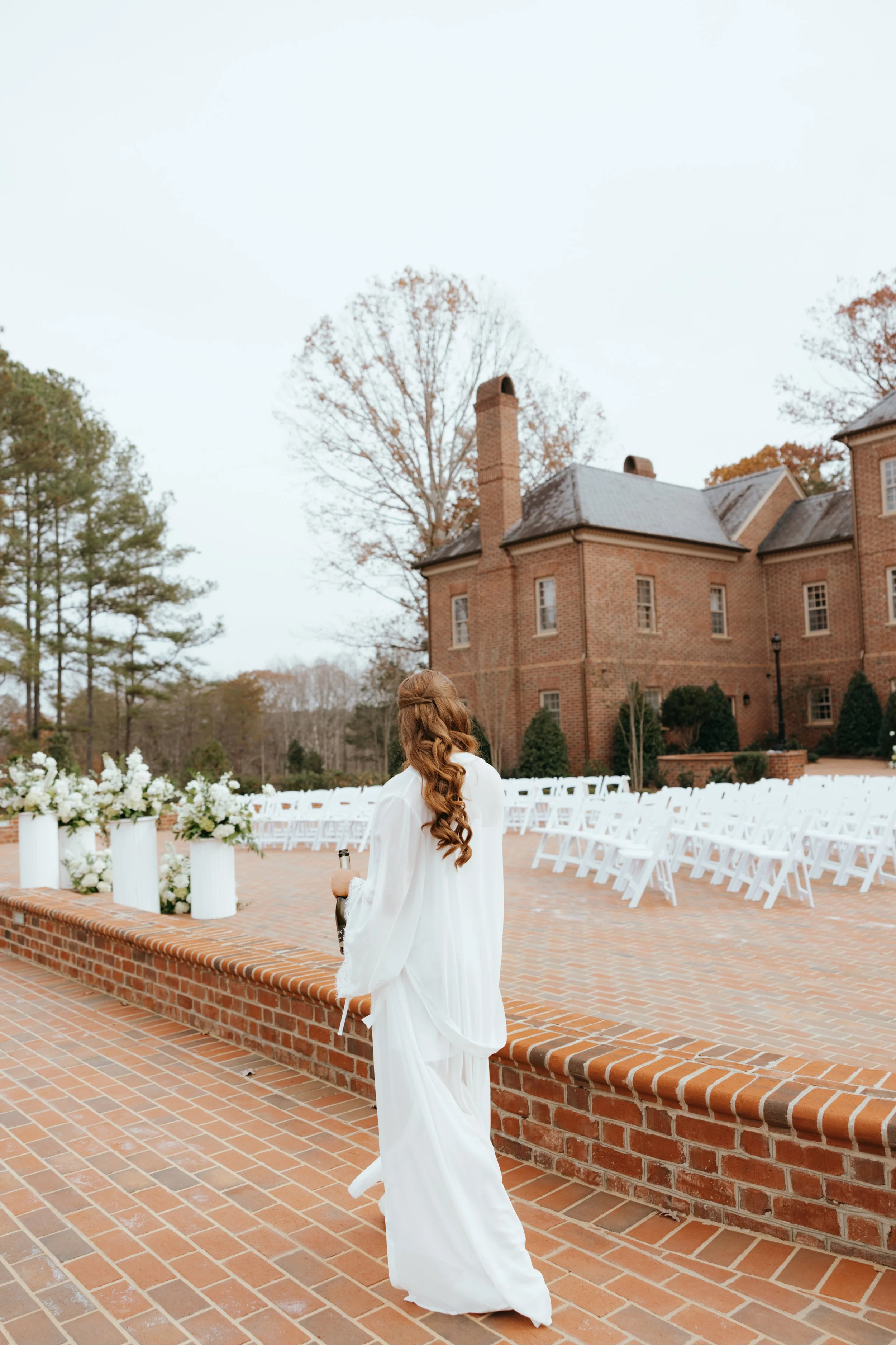 A woman in a white dress walking on a brick patio during a wedding or outdoor event, with a historic brick mansion and many white chairs arranged for an outdoor ceremony, surrounded by trees.