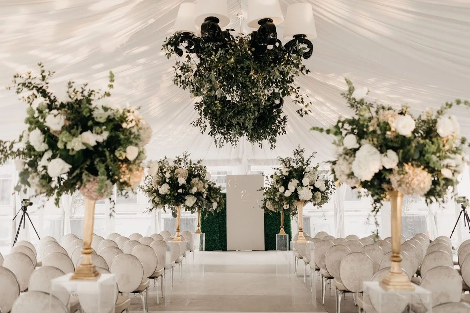 Elegant wedding ceremony setup in a white tent with rows of chairs, large floral arrangements on gold stands, and a white backdrop at the altar with greenery accents.