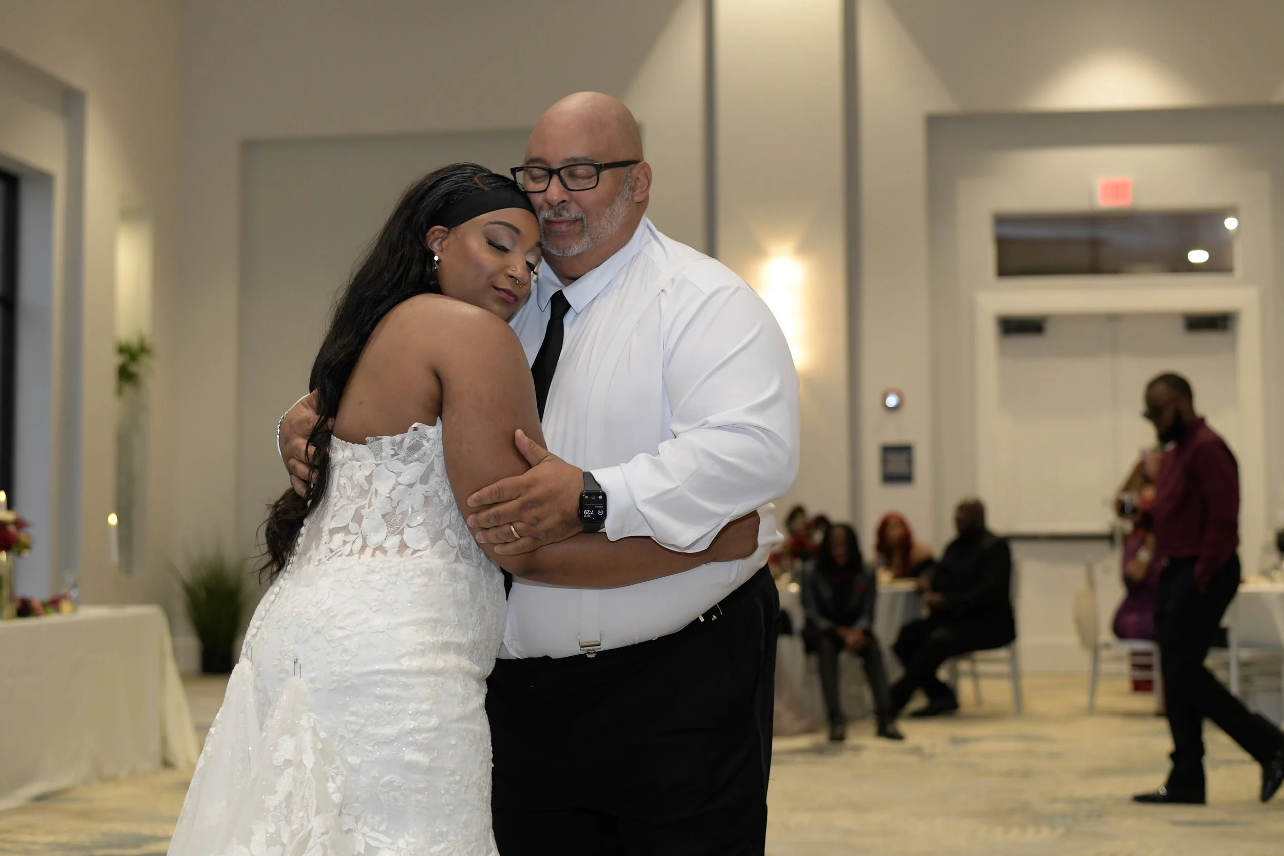 A woman in a white wedding dress and a man in a white shirt and black tie hugging and sharing a moment at a wedding reception with other guests in the background.
